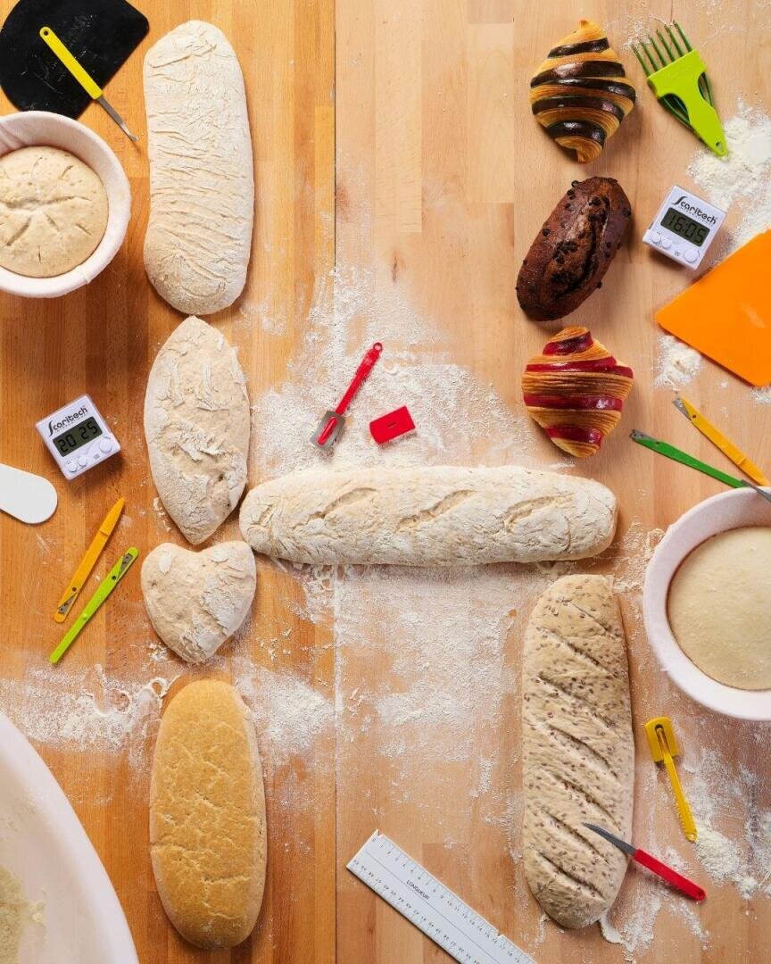 Ein Holztisch ist mit rohem und gebackenem Brot in verschiedenen Formen bedeckt. Backutensilien, Küchenwecker, Lineale und Mehl liegen verstreut herum. Manche Brote liegen in Gärschalen, andere stehen auf dem Tisch, bereit zum Backen oder bereits gebacken.