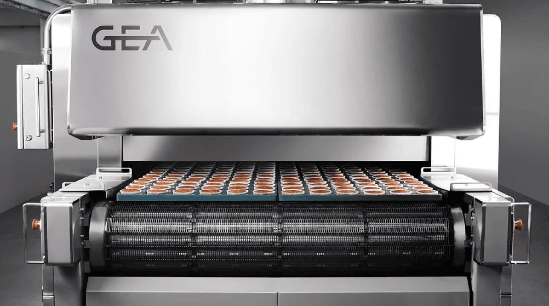 Eine industrielle Lebensmittelverarbeitungsmaschine aus Edelstahl mit der Marke GEA bewegt ein Förderband, das mit Reihen von kleinen, runden Backwaren oder Desserts gefüllt ist.