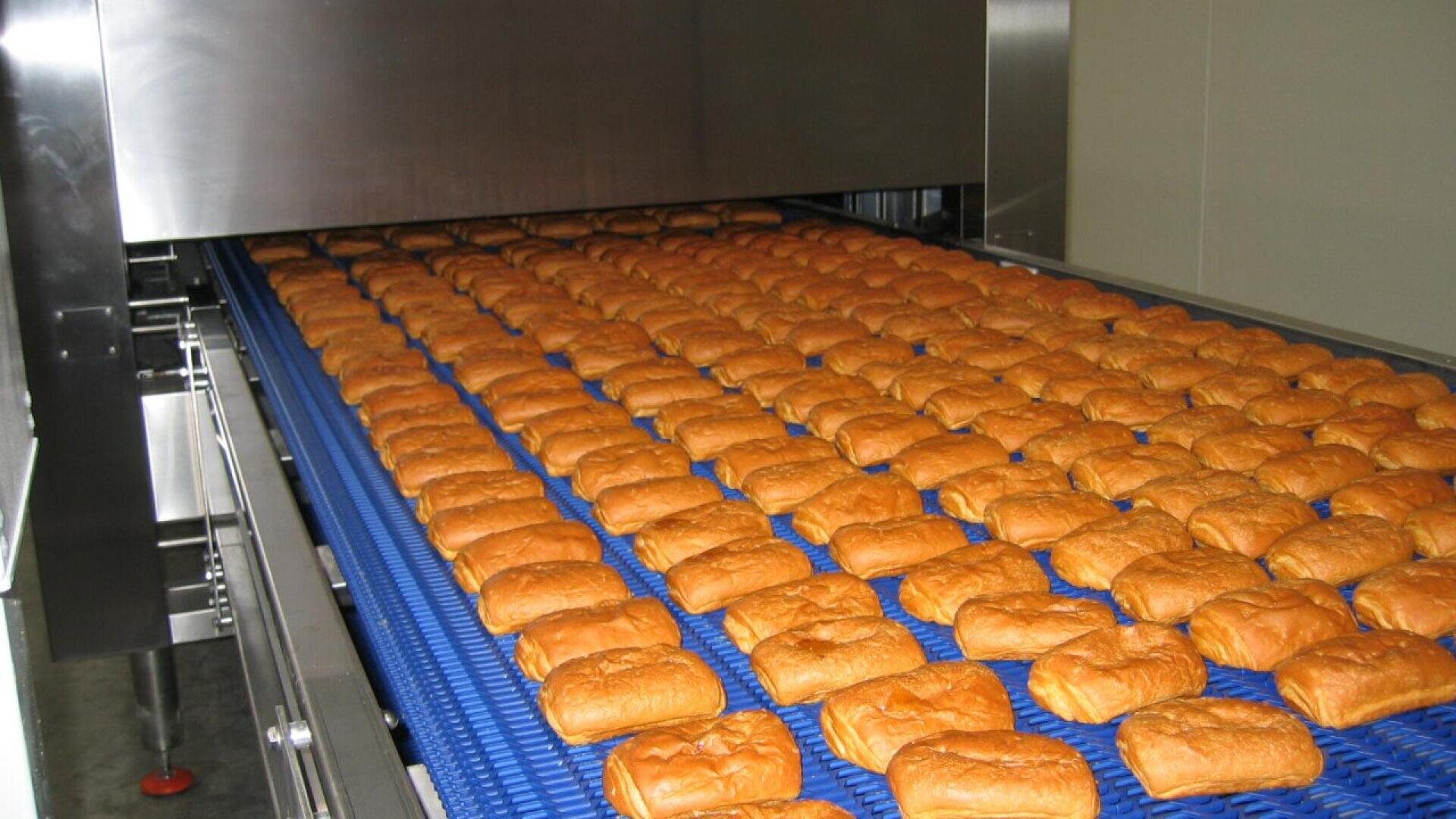 Reihen von goldbraunen Brötchen bewegen sich auf einem blauen Förderband in einer gewerblichen Bäckerei und durchlaufen industrielle Edelstahlgeräte zur Verarbeitung oder zum Backen.
