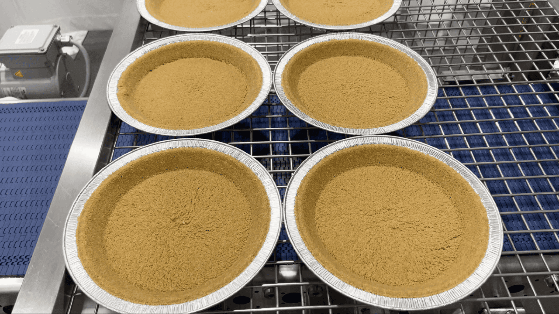 Sechs leere Kuchenkrusten in Folienschalen sind auf einem Metallkühlregal oder einem Förderband in einer scheinbar gewerblichen Küche oder Bäckerei angeordnet.