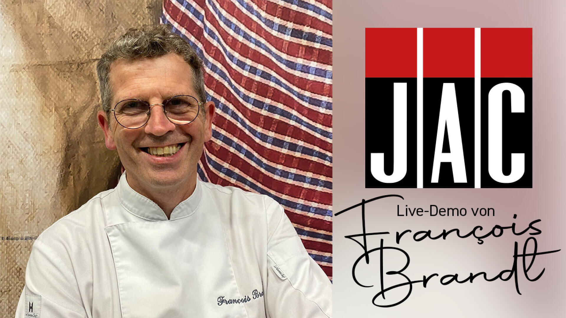 Ein lächelnder Mann in einem weißen Kochmantel steht neben einem Schild mit der Aufschrift "JAC Live-Demo von François Brandt" mit einem roten, schwarzen und weißen Logo. Im Hintergrund hängt ein gestreiftes Tuch.