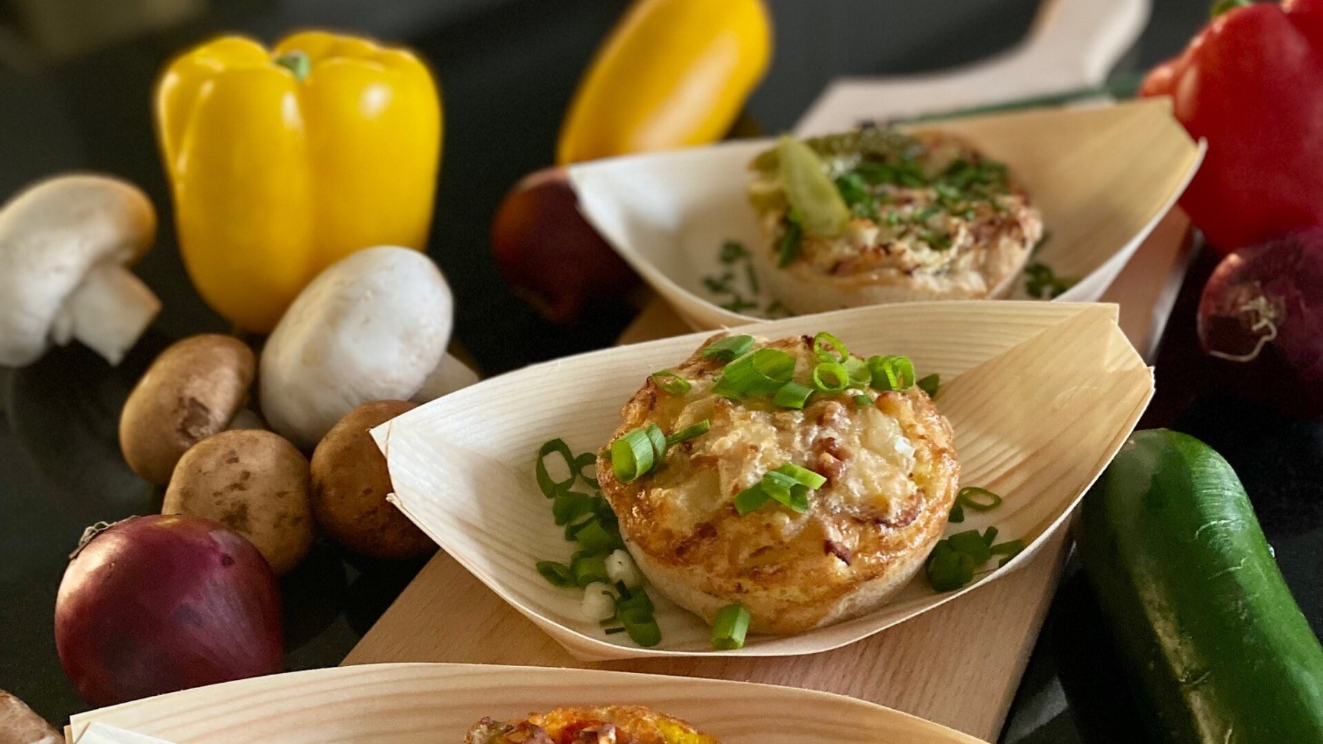 Drei einzelne Eiermuffins, garniert mit gehackten grünen Zwiebeln, werden in Holztabletts serviert, umgeben von frischem Gemüse, darunter Pilze, gelbe Paprika, rote Zwiebeln und eine Zucchini auf einer dunklen Fläche.