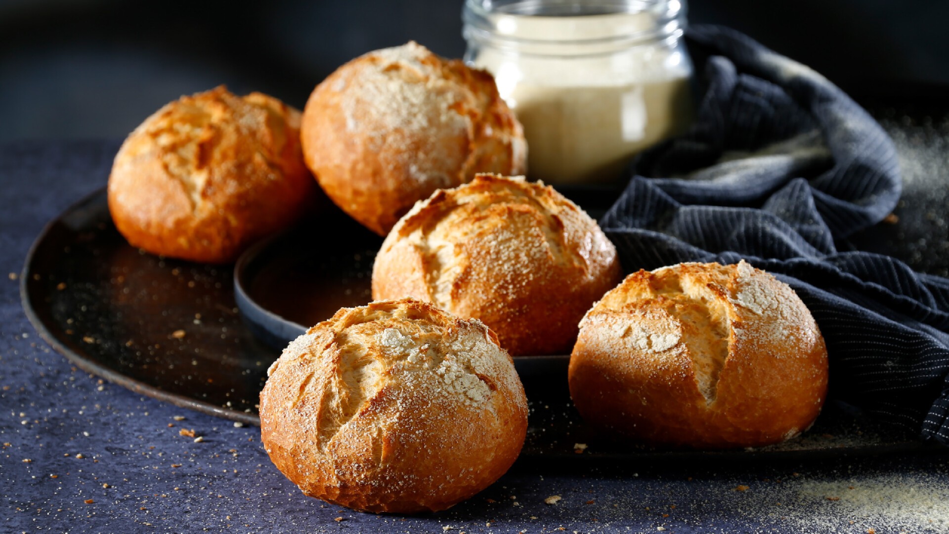Five round, golden-brown bread rolls dusted with flour are arranged on a dark tray. A glass jar of flour and a dark cloth are in the background. The scene is softly lit and rustic.