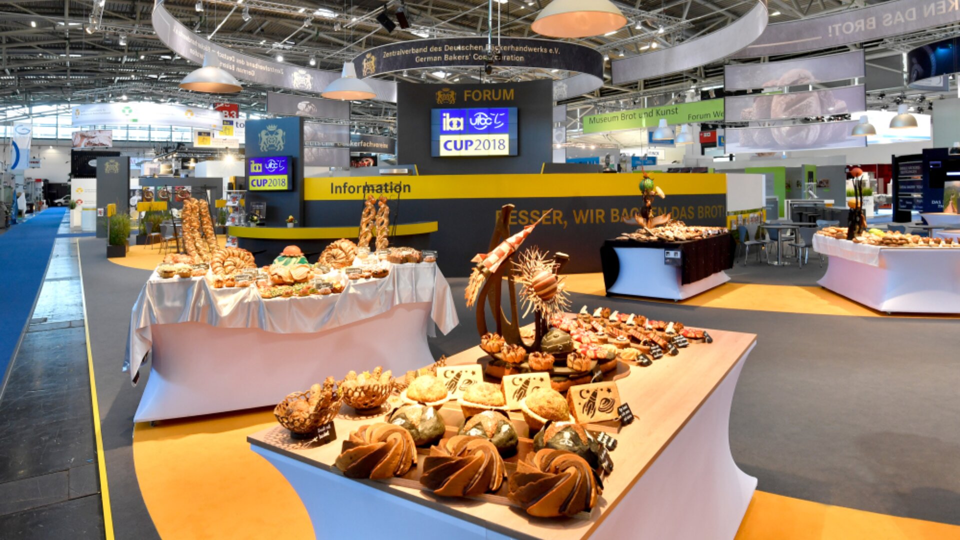 Auf einer Bäckereifachmesse sind auf Tischen verschiedene dekorative Brotsorten und Gebäckstücke ausgestellt. Im Hintergrund sind Stände und Informationsschilder zu sehen, die hell beleuchtet sind und in einer geräumigen Ausstellungshalle stehen.