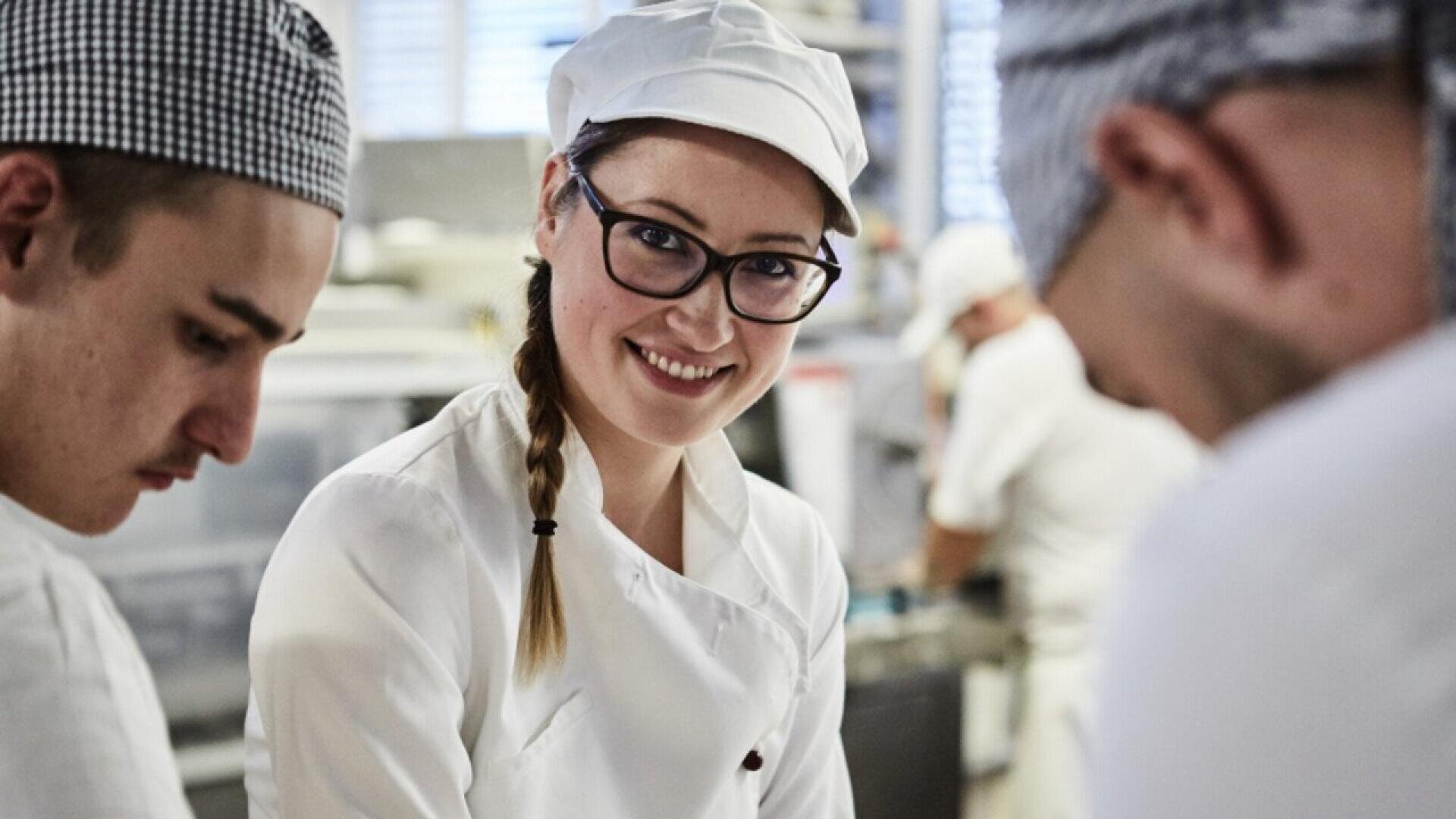 A smiling chef wearing glasses and a white uniform stands between two other chefs in a busy kitchen, looking at the camera. The background shows kitchen equipment and staff at work.