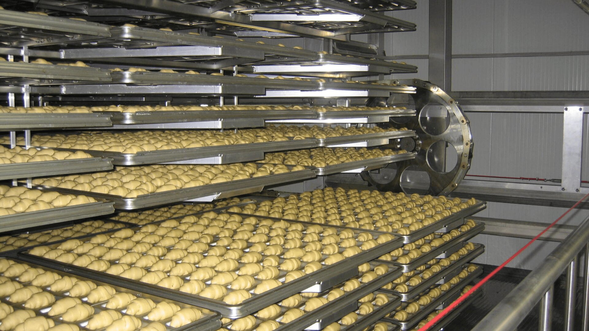 Multiple metal racks holding trays filled with rows of unbaked dough pieces inside an industrial baking or proofing room.