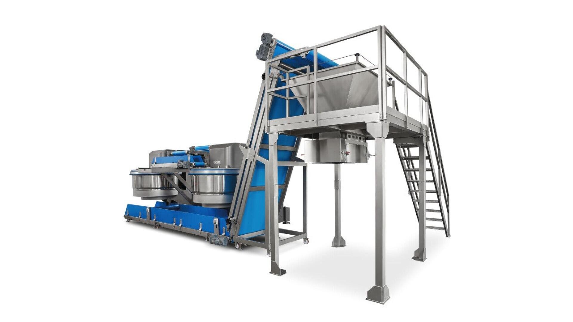Große industrielle Lebensmittelverarbeitungsmaschine mit blauen und silbernen Metallteilen, darunter ein Fördersystem, zwei runde Behälter und eine erhöhte Plattform mit Treppe und Geländer für den Zugang des Bedieners.
