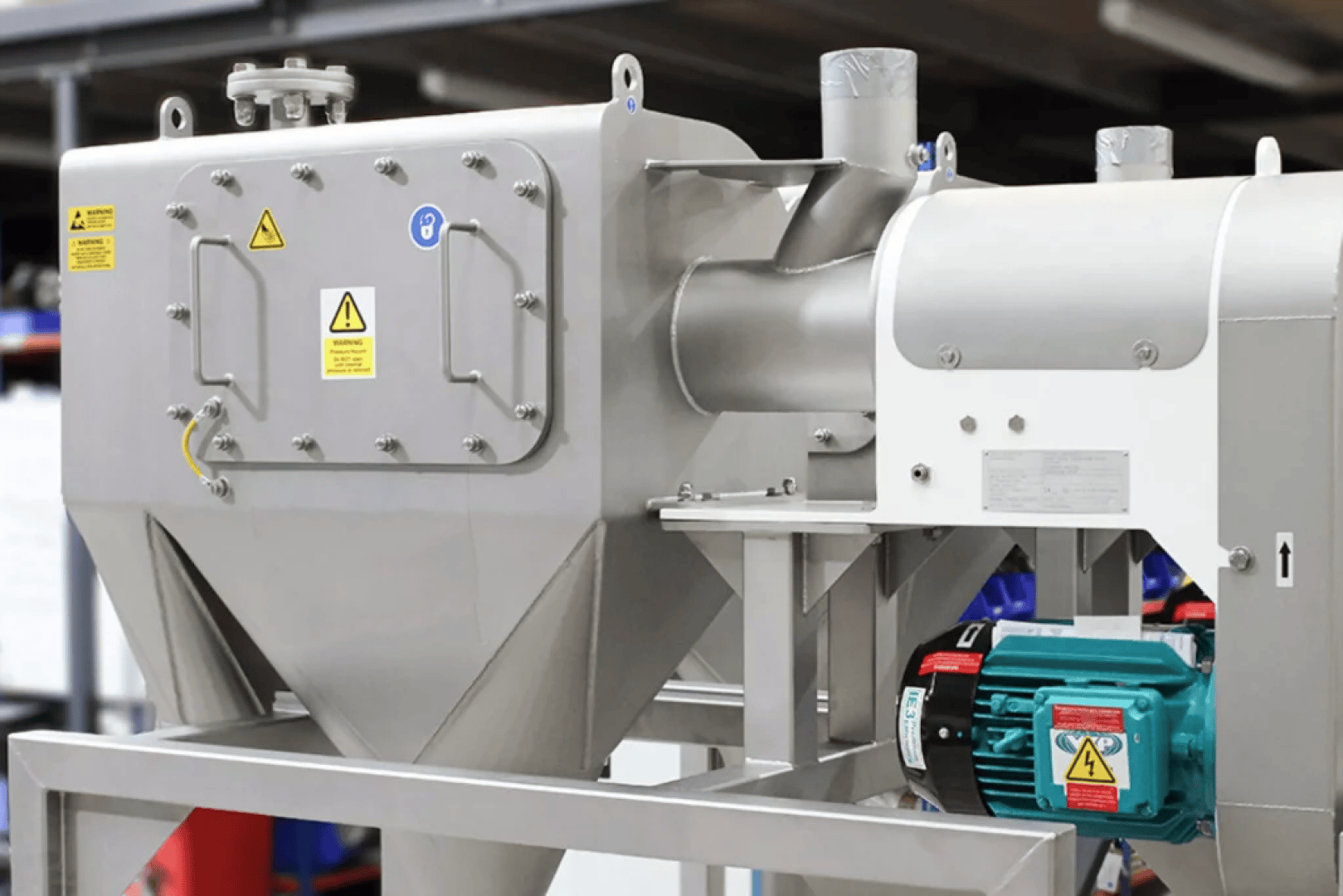 Industrielle Maschinen aus rostfreiem Stahl mit Warnschildern, montiert auf einem Rahmen. An einer Seite ist ein grüner Elektromotor angebracht. Die Ausrüstung scheint für die Verarbeitung oder Herstellung in einer industriellen Umgebung verwendet zu werden.