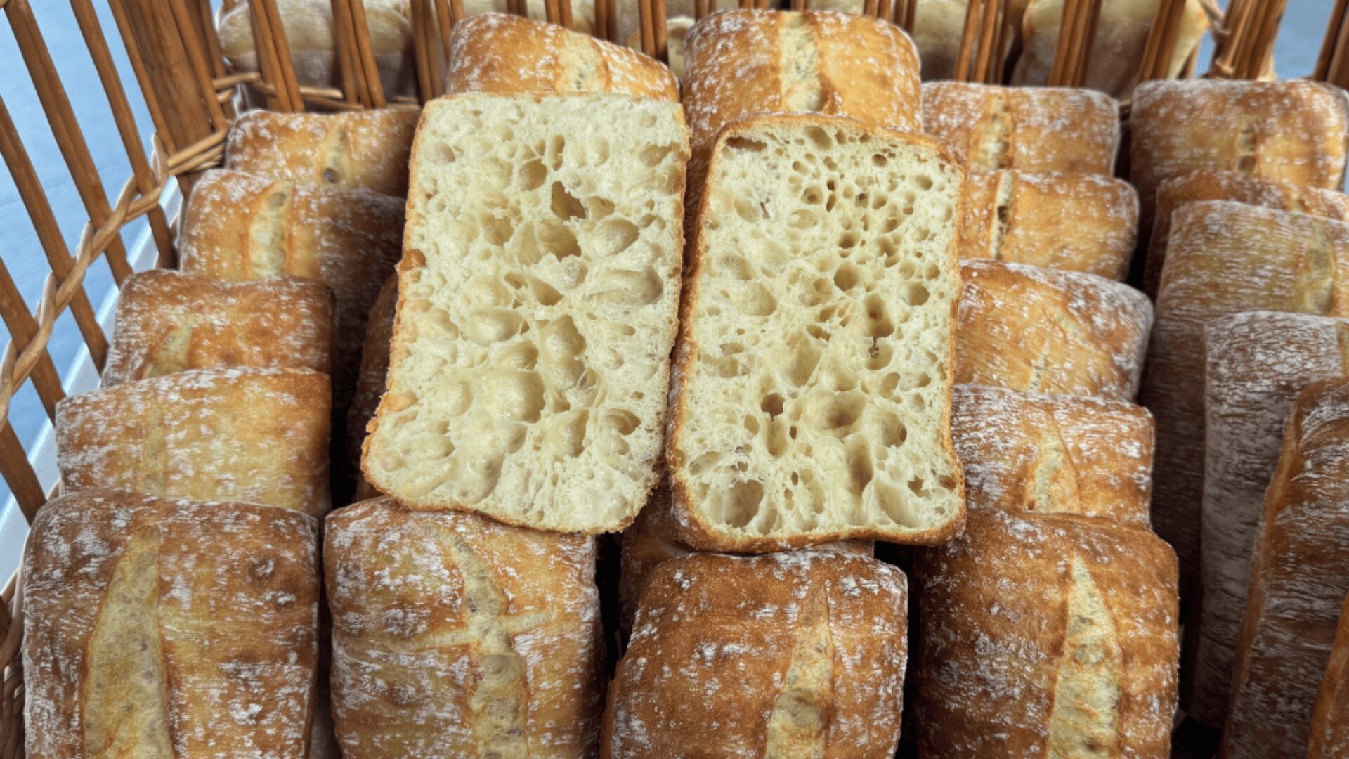 A basket filled with rectangular ciabatta rolls, two of which are cut open to show their airy, hole-filled crumb structure and golden-brown crust.