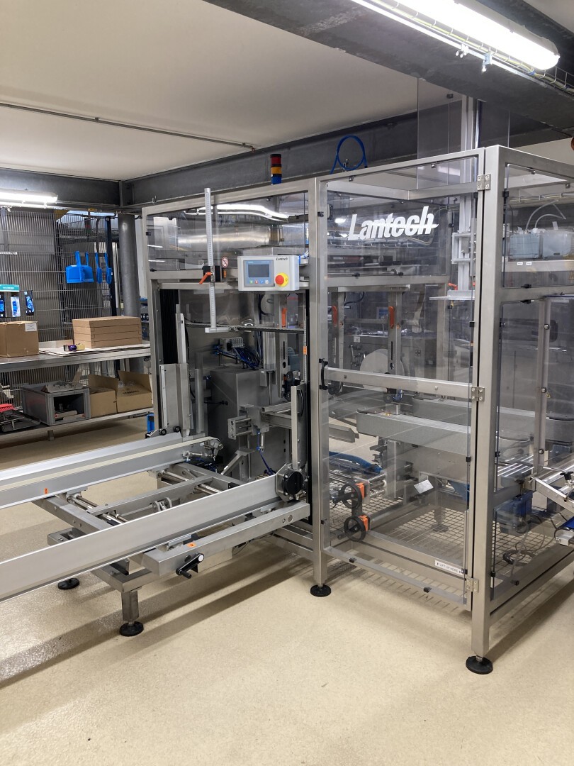 Eine Lamtech-Verpackungsmaschine aus Edelstahl steht in einem gut beleuchteten Industrieraum, umgeben von Förderbändern, Bedienfeldern und Regalen mit Kisten und Geräten.