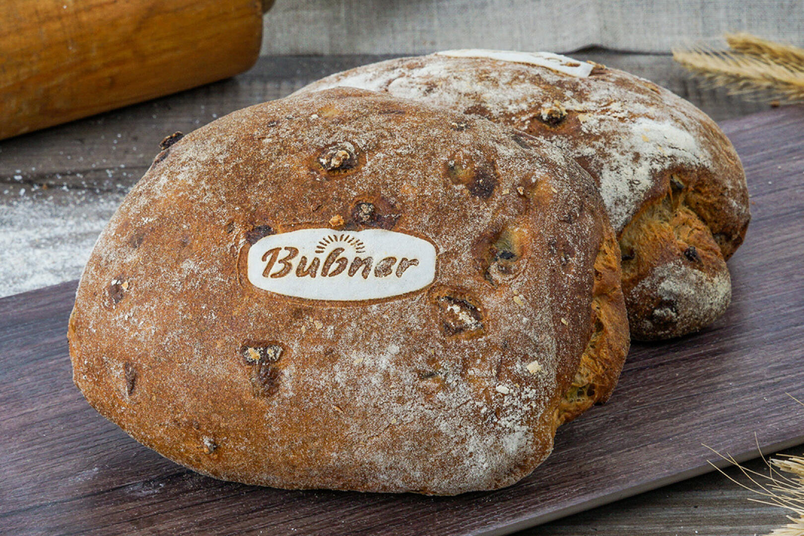 Zwei rustikale, mit Mehl bestäubte Brote, eines davon mit einem Etikett mit der Aufschrift Bubner, liegen auf einer hölzernen Fläche mit Weizen und einem Nudelholz im Hintergrund.