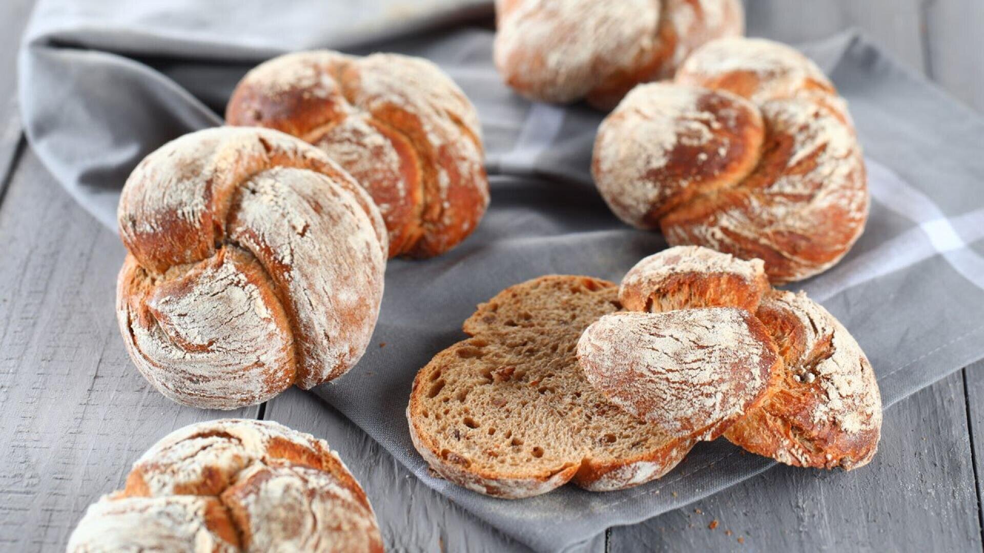 Round loaves of rustic bread with a dusting of flour are arranged on a gray cloth atop a wooden table. One loaf is sliced open, showing the soft, brown interior.