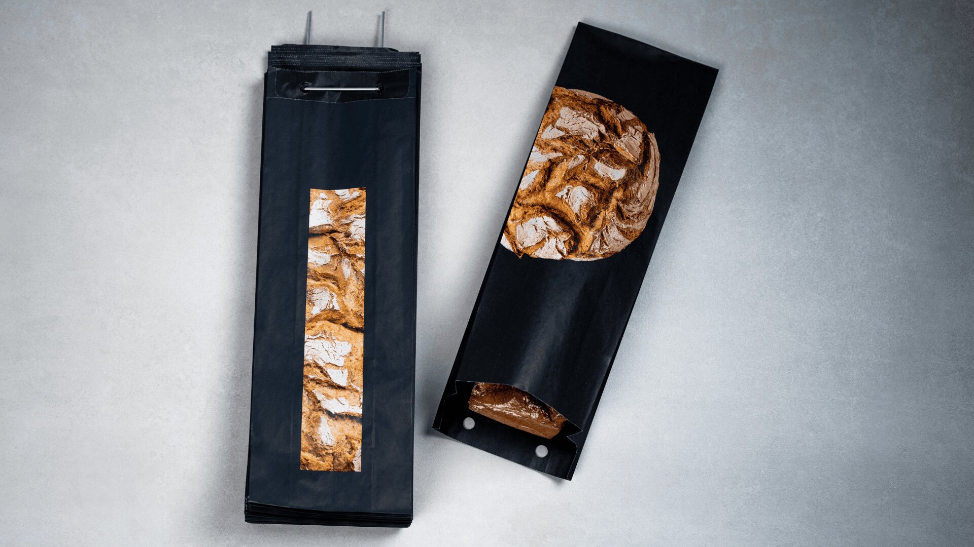 Auf einer hellgrauen Fläche liegen zwei schwarze Papiertüten mit einem durchsichtigen Fenster, in dem sich ein rustikaler Laib Brot befindet. Eine Tüte ist mit anderen gestapelt, die andere liegt daneben, teilweise geöffnet.