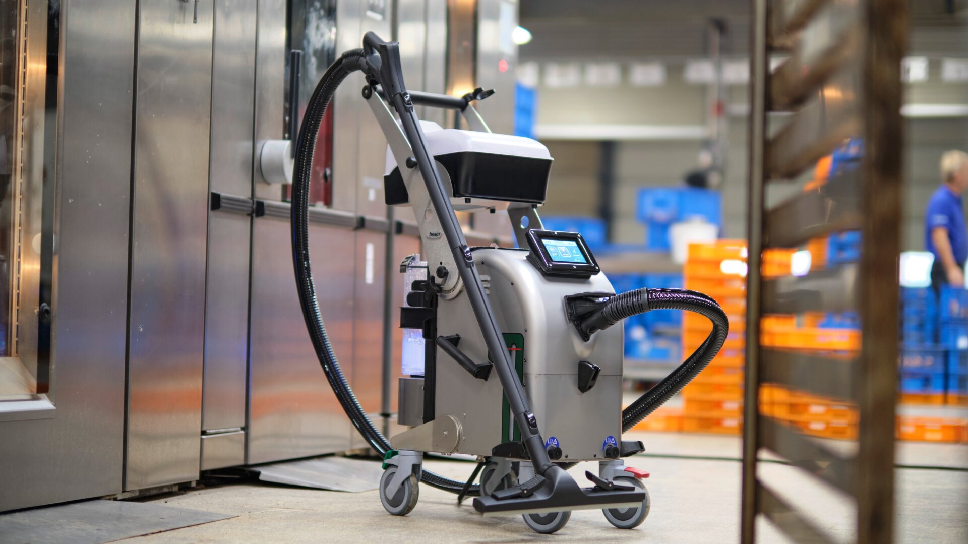 Ein Reinigungsroboter mit Schläuchen und einem Touchscreen steht auf Rädern in einer Industrieanlage in der Nähe eines großen Metalltors, mit verschwommenen Regalen und orangefarbenen Kisten im Hintergrund.