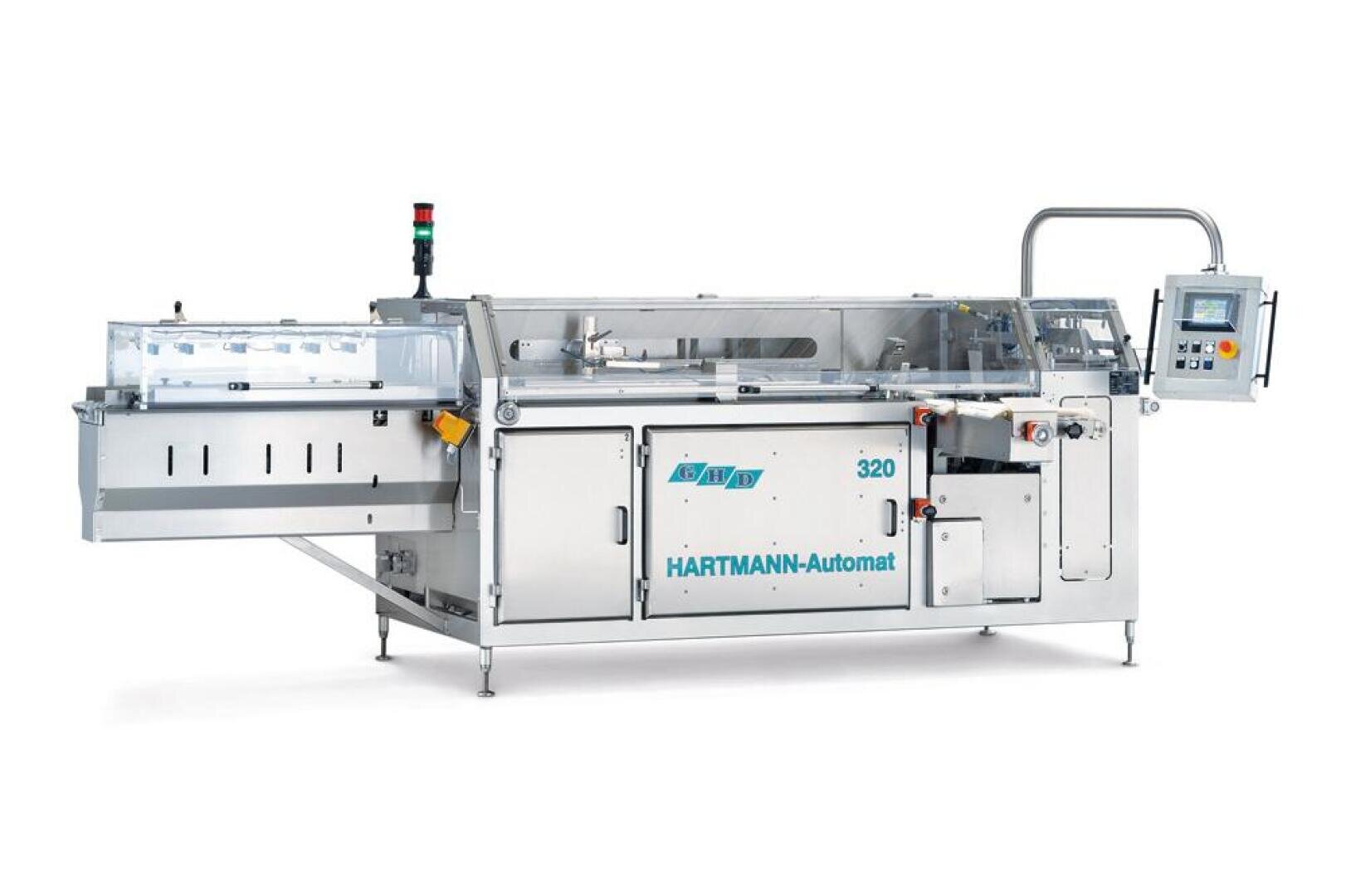 A stainless steel Hartmann-Automat 320 packaging machine with a transparent cover, control panel, emergency stop button, and various conveyor sections, designed for automated industrial packaging.