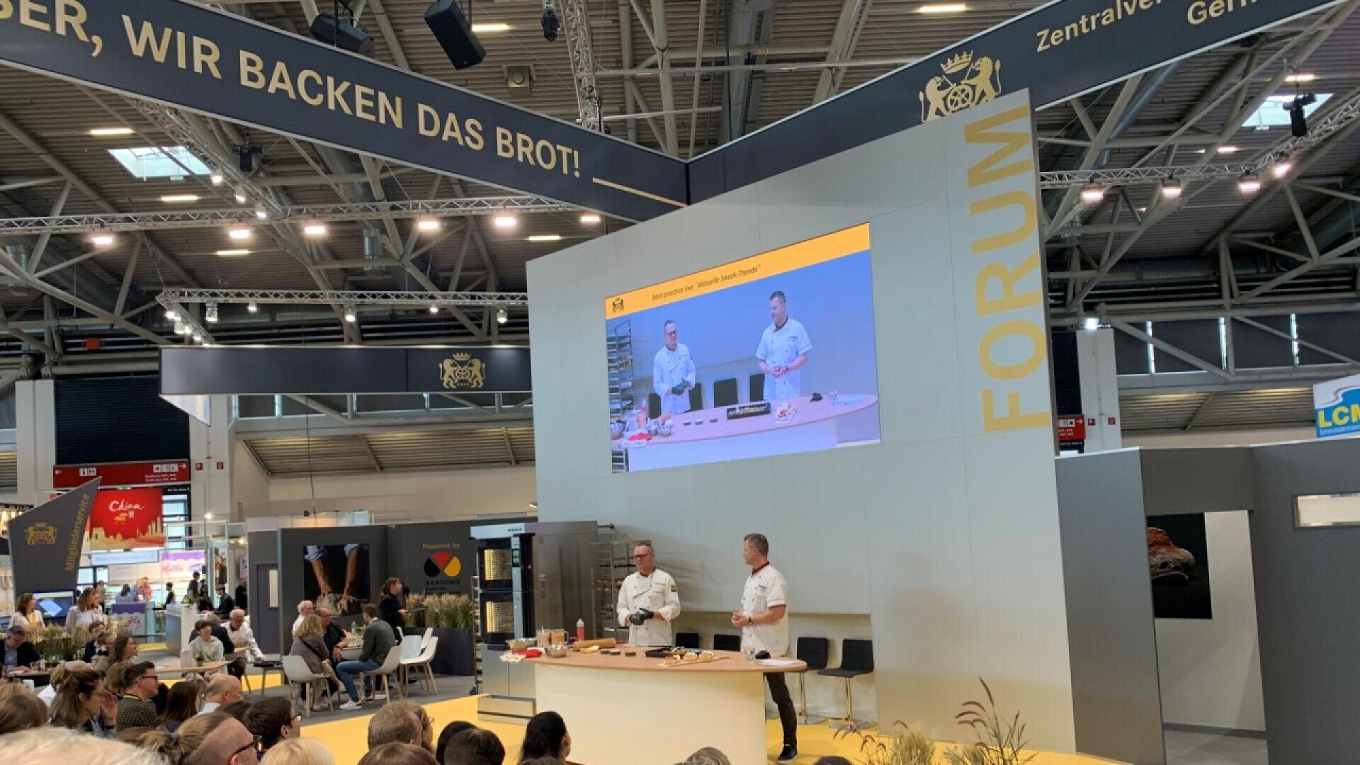 Two men in white chef coats host a baking demonstration on a stage at a large indoor event, with a crowd seated in front of them and a large screen displaying their actions above the stage.