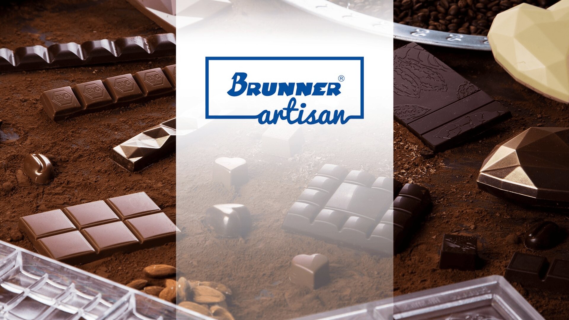 Verschiedene handwerklich hergestellte Pralinen in unterschiedlichen Formen und Größen sind auf einer mit Kakao bestäubten Oberfläche angeordnet, wobei das Brunner-Handwerkslogo auf einem weißen, durchsichtigen Banner zentriert ist.