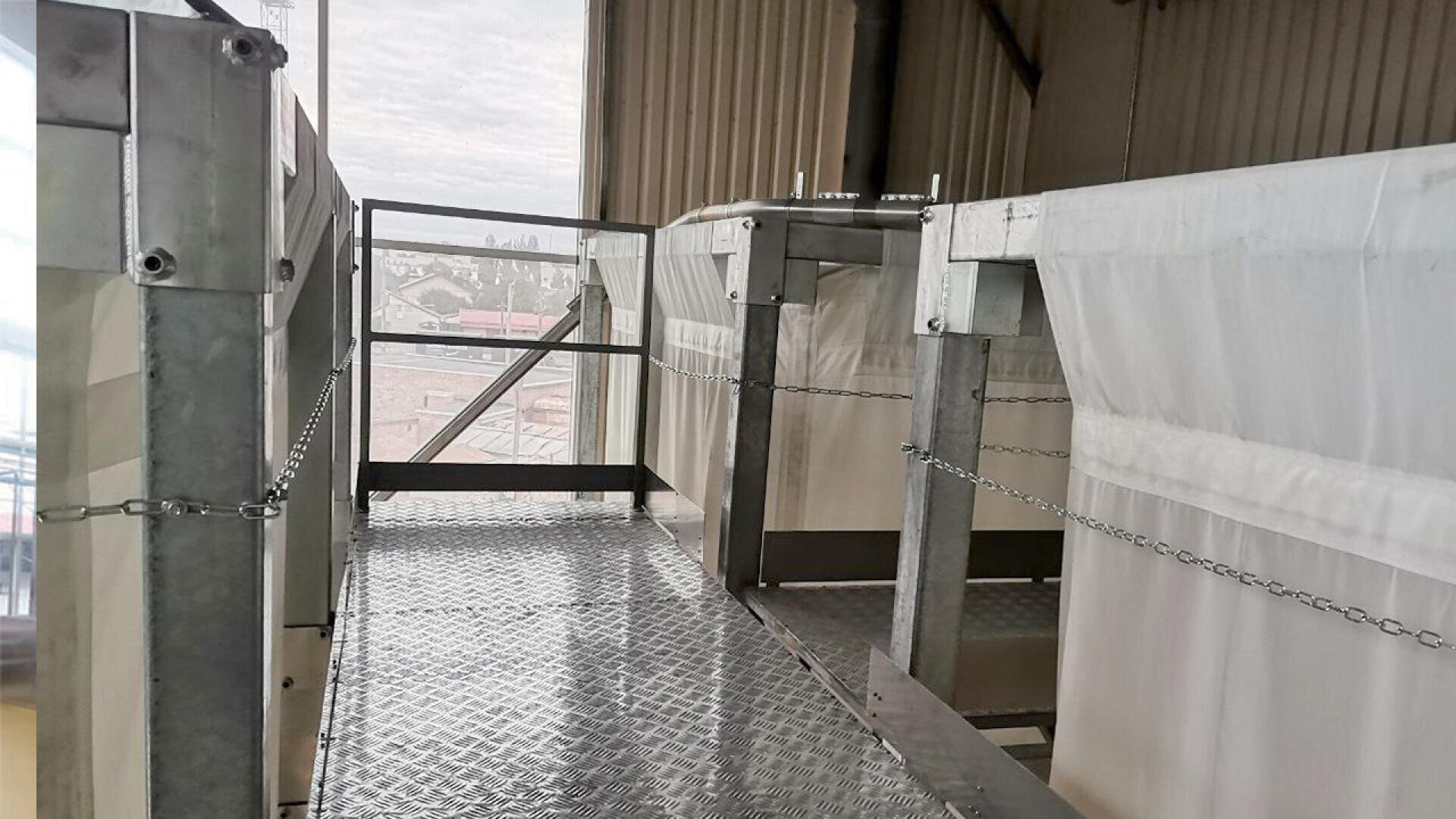 Industrieller Metallsteg mit Gitterboden und Kettenabsperrungen, im Innenbereich zwischen zwei großen Strukturen mit weißen Abdeckungen, neben einem Fenster mit Blick auf die Gebäude draußen.