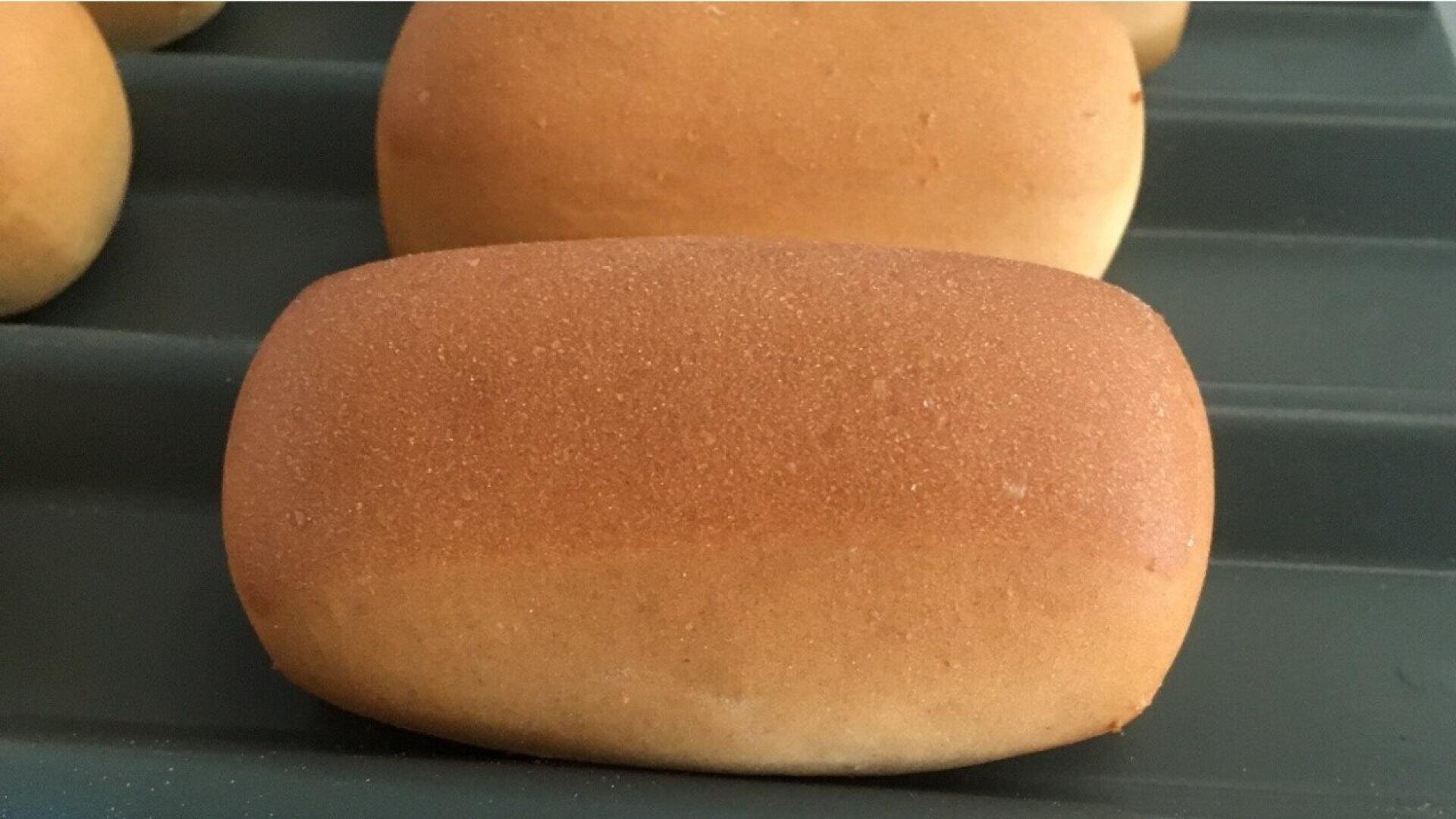 Nahaufnahme eines goldbraunen rechteckigen Brötchens, das auf einem Backblech liegt, mit anderen ähnlichen Brötchen im Hintergrund.