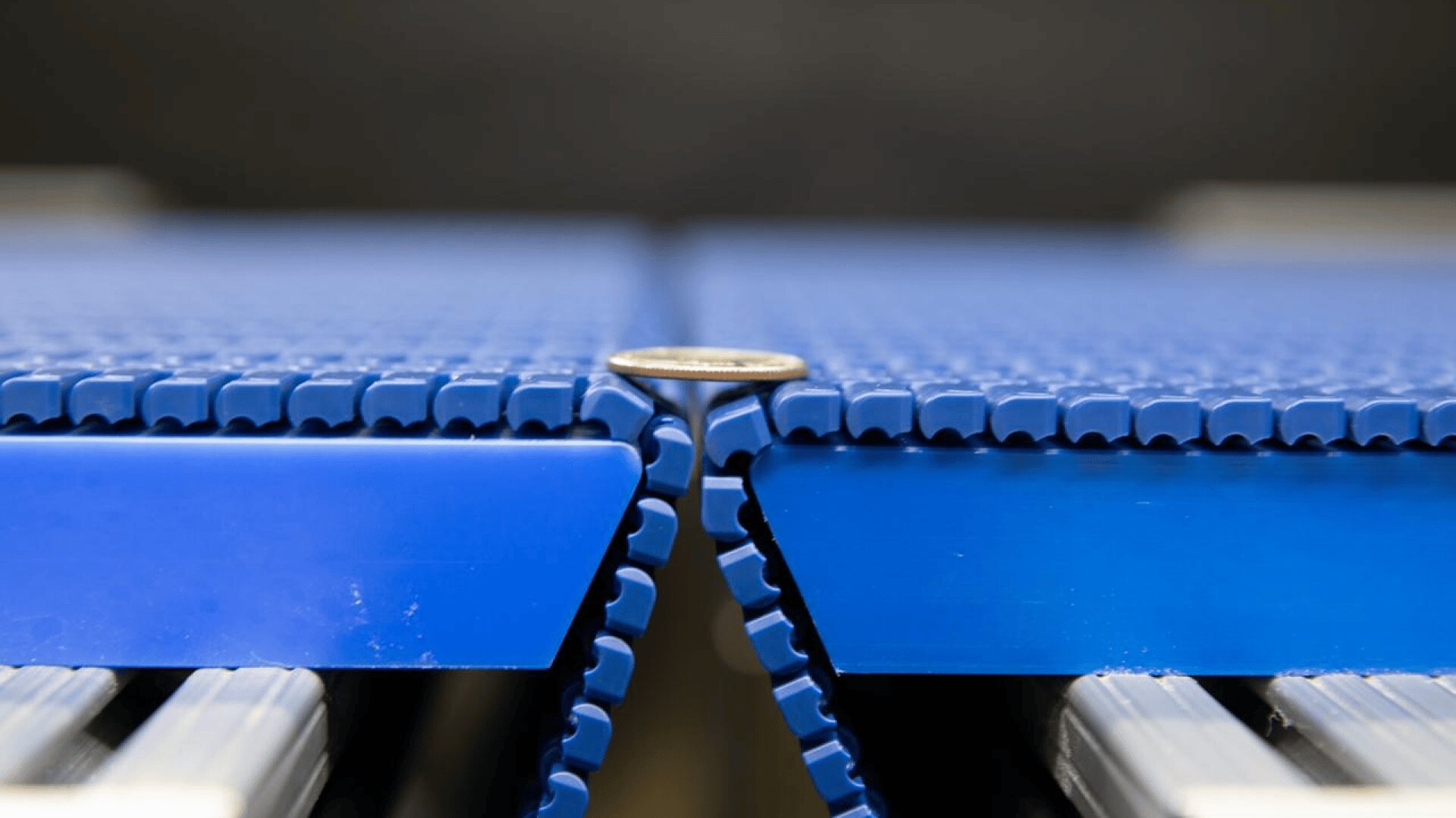 A coin stands upright between two blue conveyor belts with interlocking ridges, highlighting the narrow gap between the belt sections.
