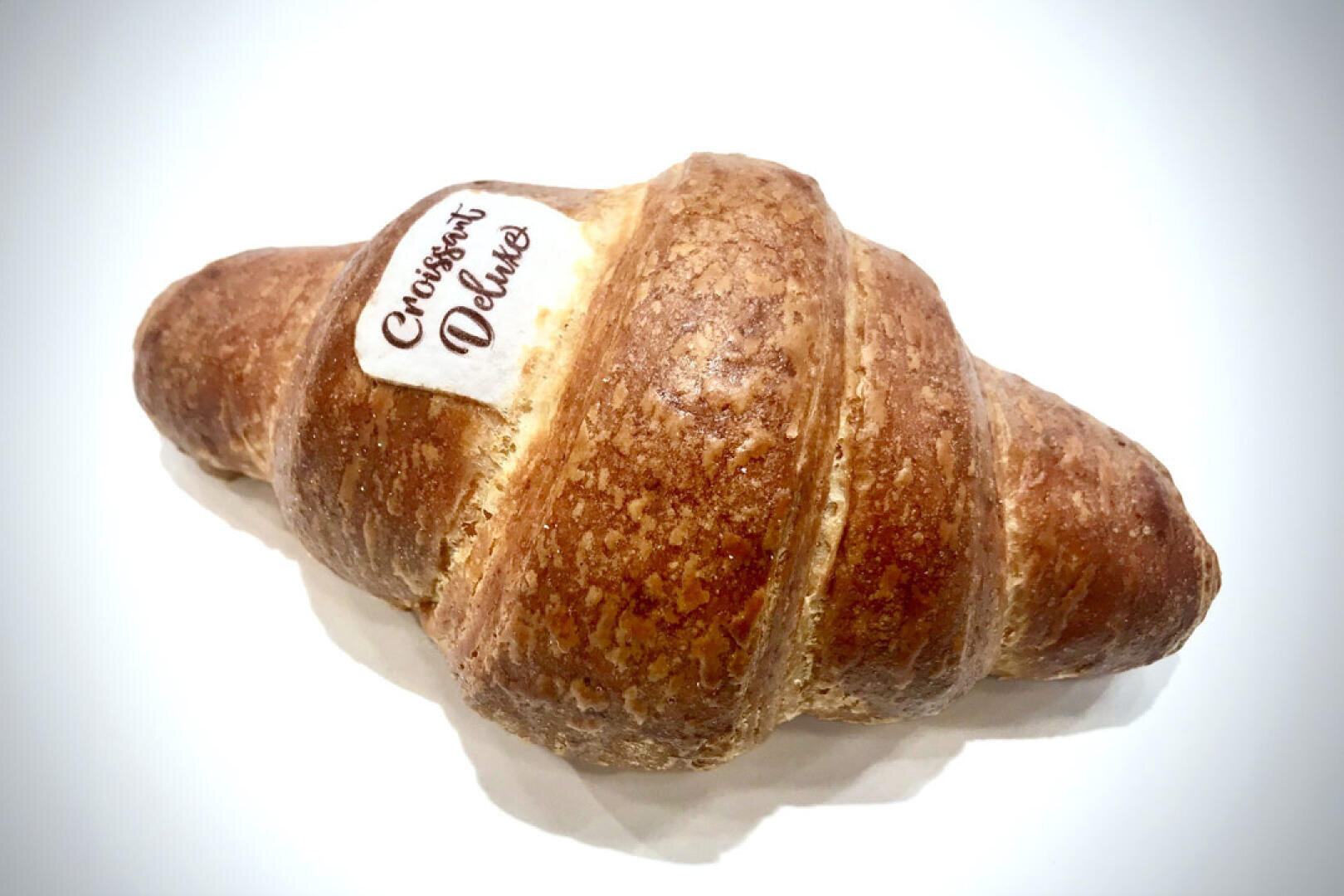 Ein goldbraunes Croissant mit einem kleinen Etikett auf der Oberseite, auf dem Croissant Deluxe steht, fotografiert auf weißem Hintergrund.