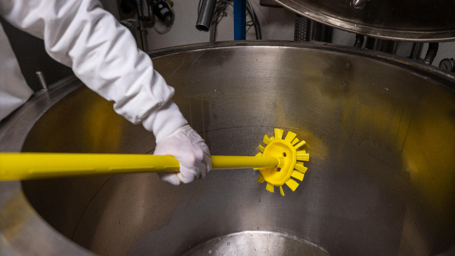Eine Person mit weißen Handschuhen reinigt mit einer gelben Bürste mit langem Stiel das Innere eines großen Industrietanks aus Edelstahl.