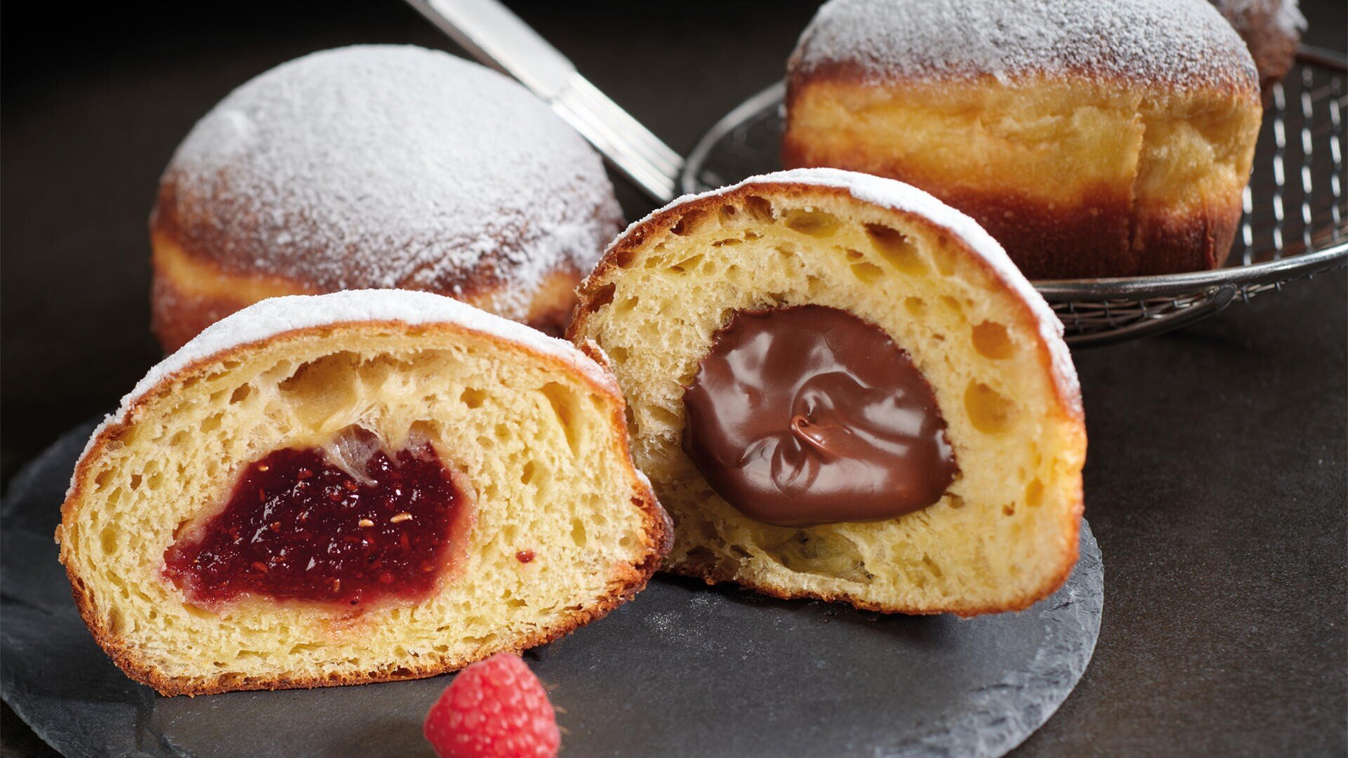 Zwei runde, mit Puderzucker bestäubte Krapfen, von denen einer aufgeschnitten ist und eine Schokoladen- und eine Himbeermarmeladenfüllung enthält, liegen auf einem Schiefertablett mit einer frischen Himbeere davor.