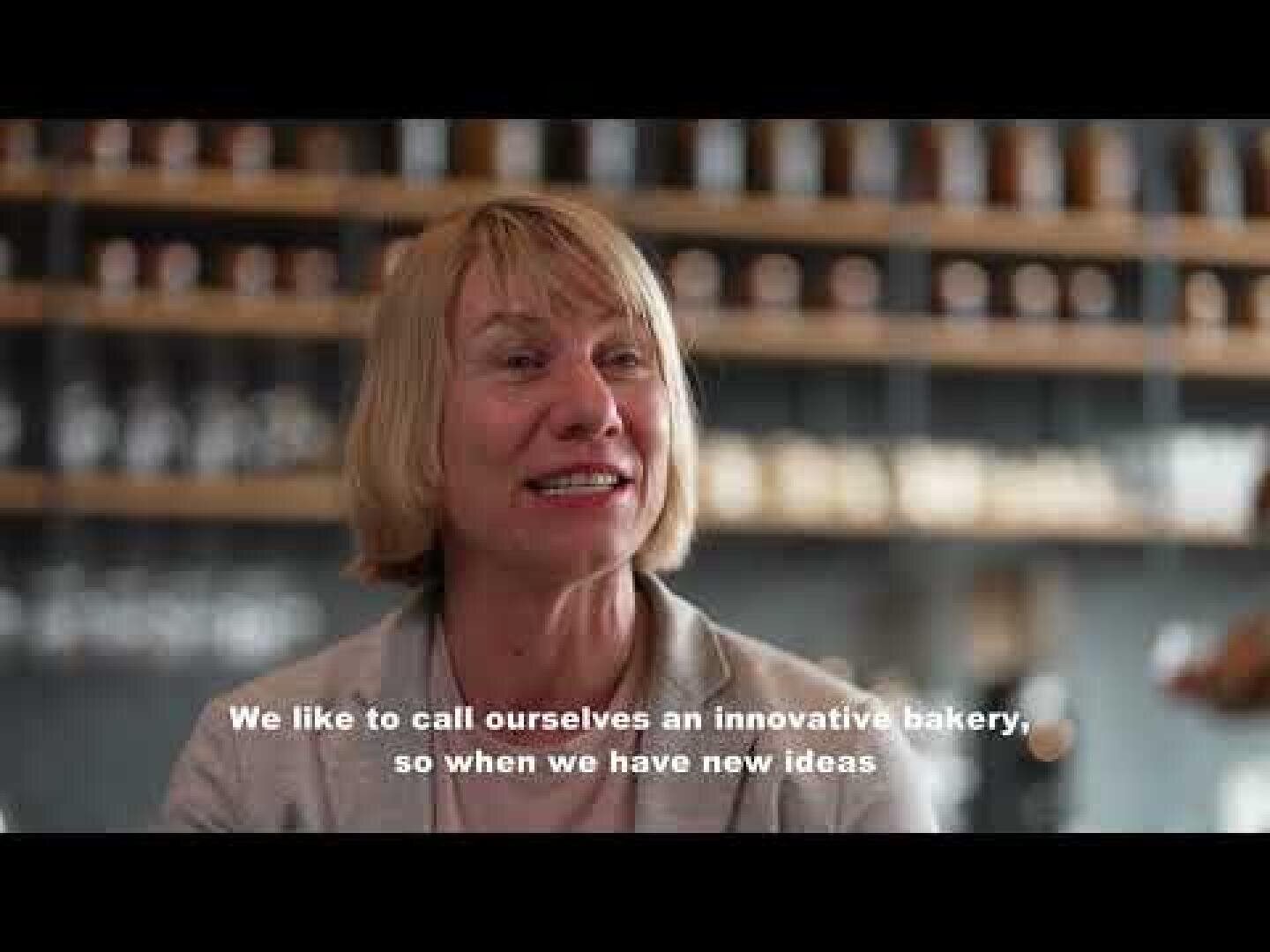 Eine lächelnde Frau mit kurzen blonden Haaren sitzt in einer modernen Bäckerei, im Hintergrund Regale mit Gläsern. In weißen Untertiteln ist zu lesen: Wir nennen uns gerne eine innovative Bäckerei, wenn wir also neue Ideen haben.
