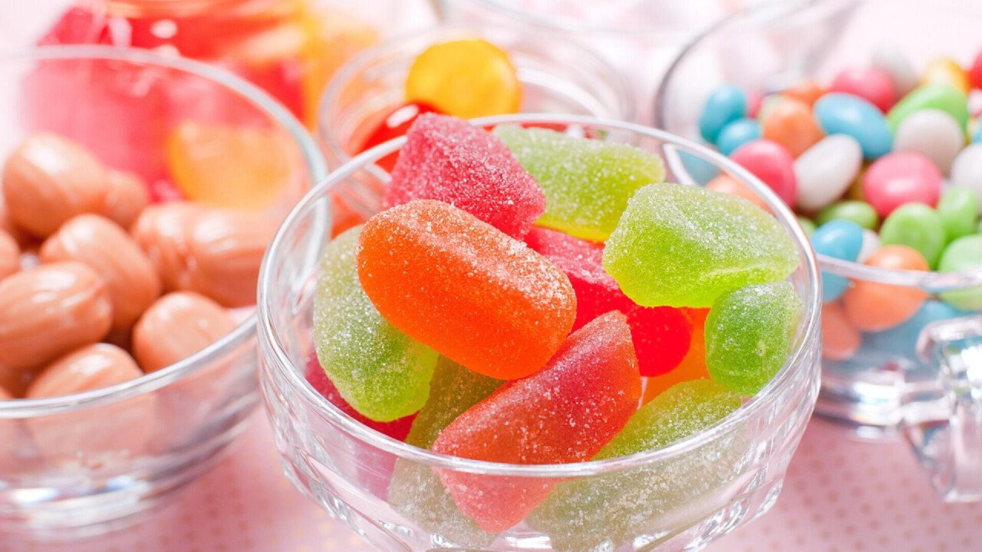 Close-up of glass bowls filled with colorful candies, including sugar-coated gummy candies, pastel round candy-coated chocolates, and shiny, oval-shaped sweets. The candies are vibrant and arranged on a light surface.