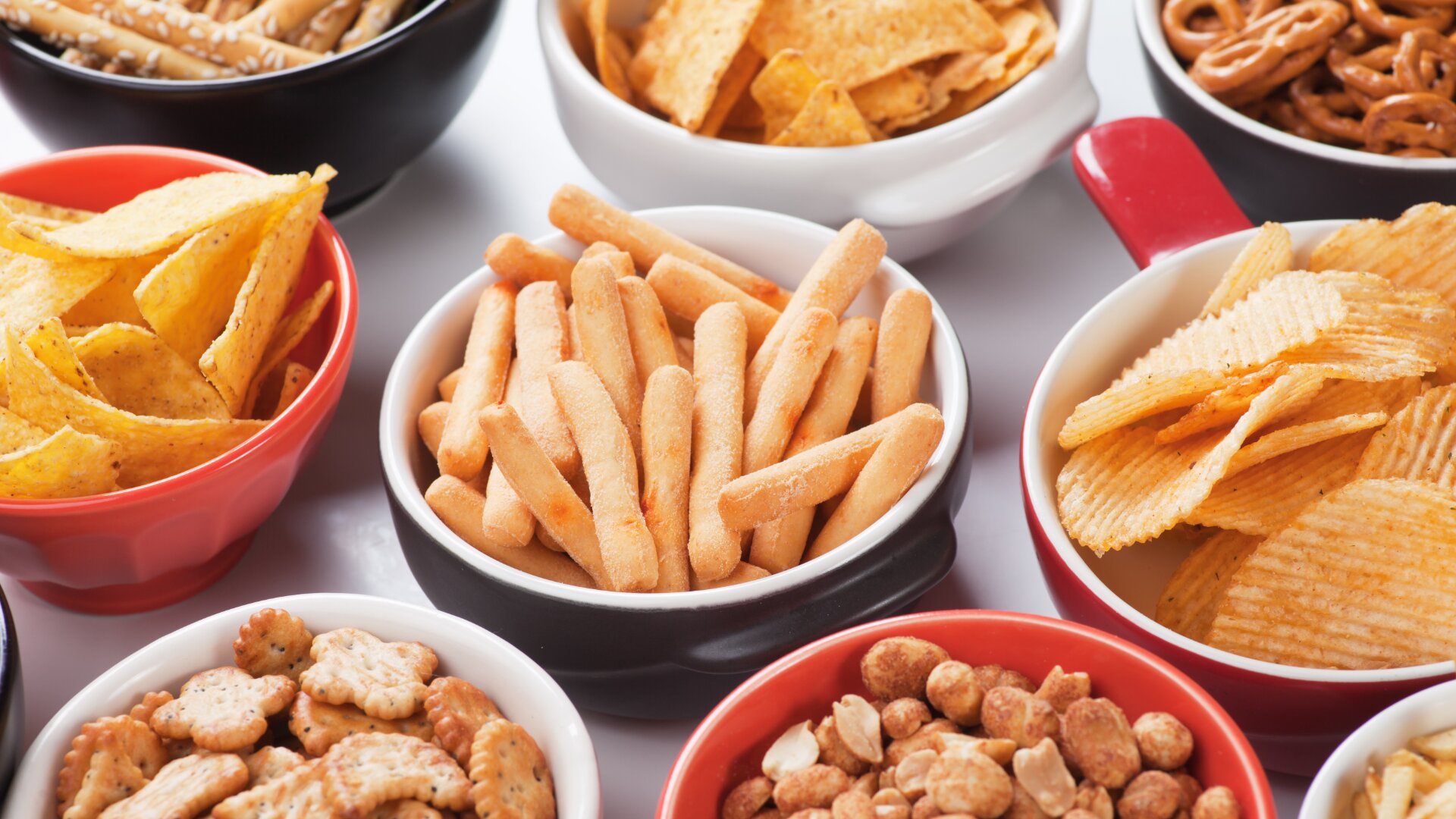 Eine Auswahl an Snacks wie Cracker, Brotstangen, Tortilla-Chips, Kartoffelchips, Brezeln und gemischte Nüsse, die in bunten Schüsseln auf einer weißen Fläche angeordnet sind.