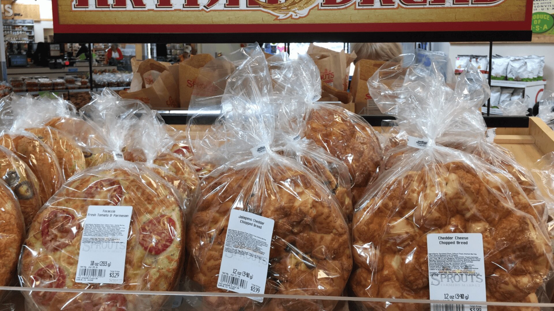 Several loaves of bread, wrapped in clear plastic, are displayed on a store shelf. The label above reads ARTISAN BREAD and the visible labels indicate tomato and parmesan as well as cheddar cheese varieties.
