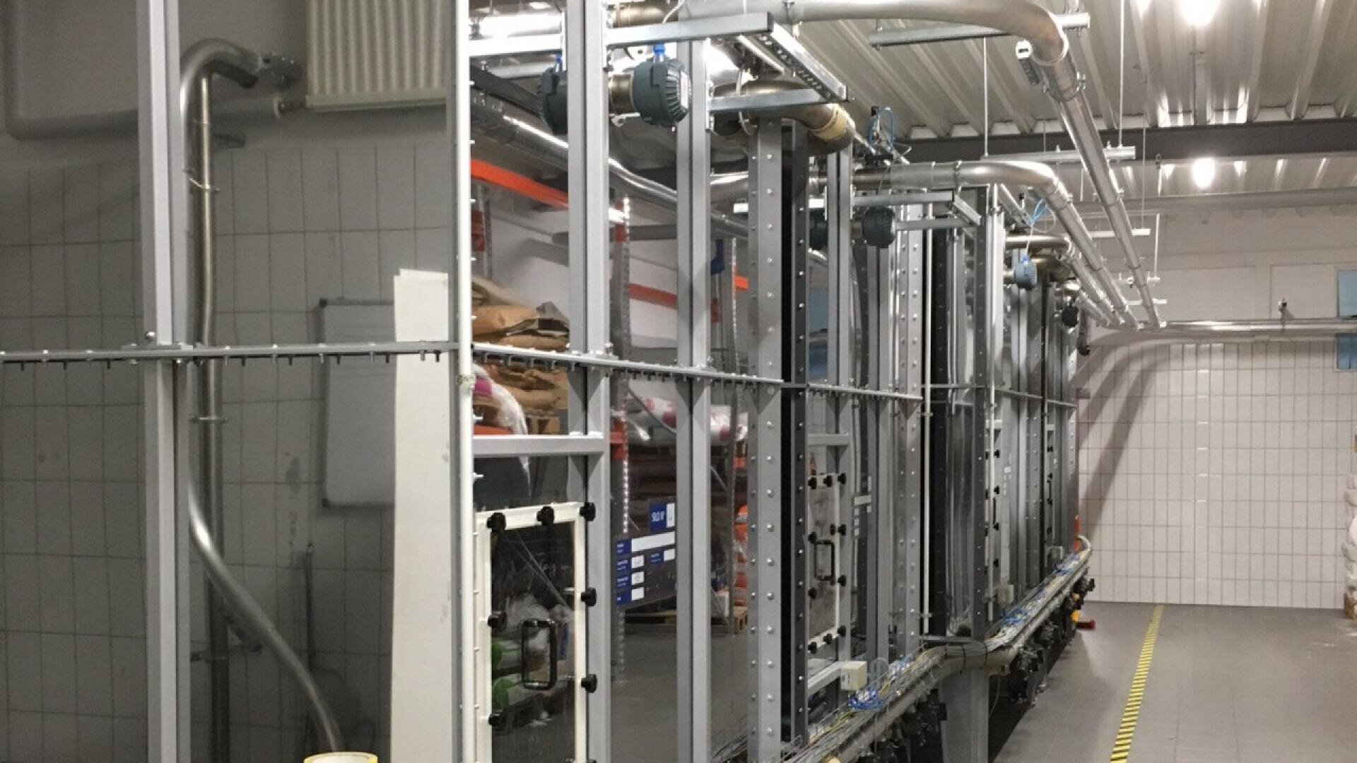 A row of industrial machines and metal pipes inside a factory with tiled walls, a metal ceiling, and caution tape marking the floor along the production line.