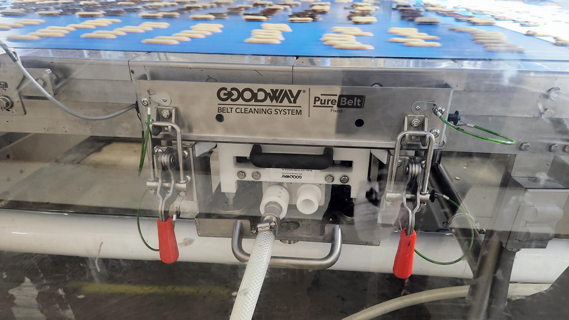 A conveyor belt cleaning system labeled Goodway PureBelt cleans a moving blue conveyor belt carrying small, assorted cookies in a food processing facility.