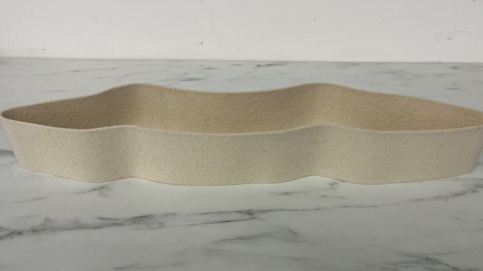 A beige, oval-shaped felt container with wavy edges sits on a white marble surface against a plain white background.