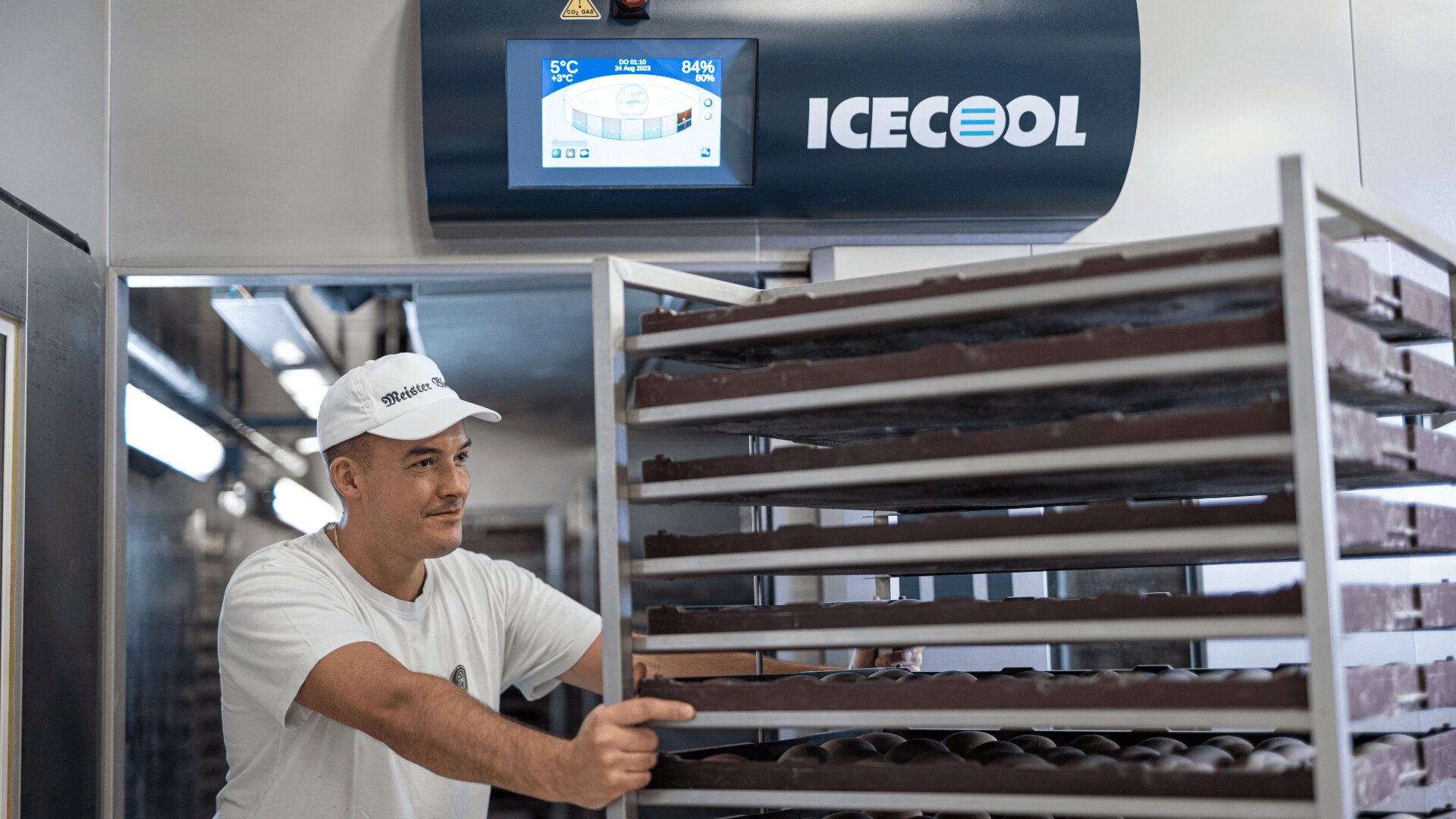 A worker in a white uniform and cap moves a rack of trays filled with chocolates inside a facility. A machine labeled ICECOOL and a digital control panel are visible in the background.