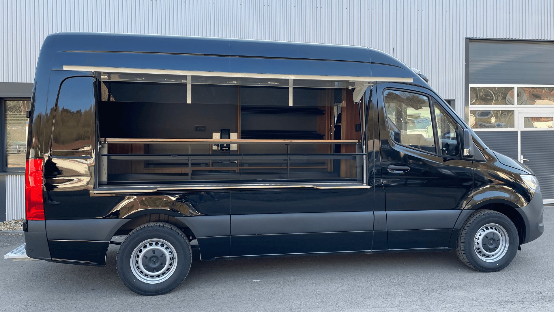 Ein eleganter schwarzer Imbisswagen mit einem großen offenen Servierfenster, geparkt auf einer gepflasterten Fläche vor einem modernen Industriegebäude. Das Innere des Lastwagens wirkt sauber und leer, bereit für den Einsatz.