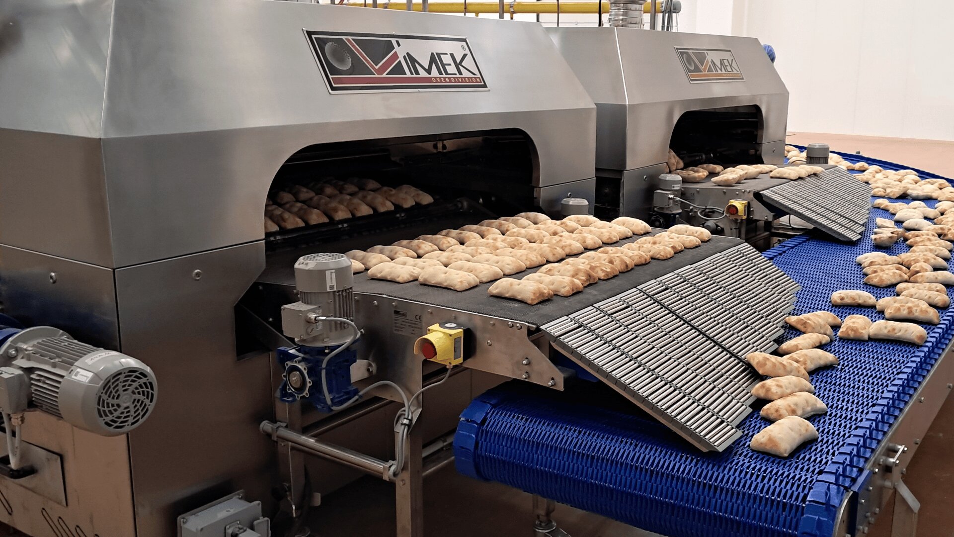 Reihen von Backwaren bewegen sich auf Förderbändern durch große industrielle Backmaschinen in einer Fabrikumgebung. Einige Backwaren verlassen die Maschinen auf blauen Förderbändern.
