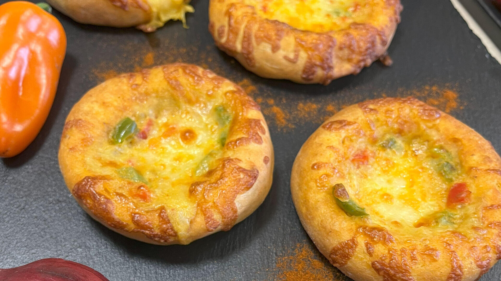 Auf einer schwarzen Fläche sind vier Mini-Käse- und Gemüsebrotschalen mit goldener, geschmolzener Käseoberfläche angeordnet, an der Seite sind eine orange Paprika und ein Hauch von Rot zu sehen.