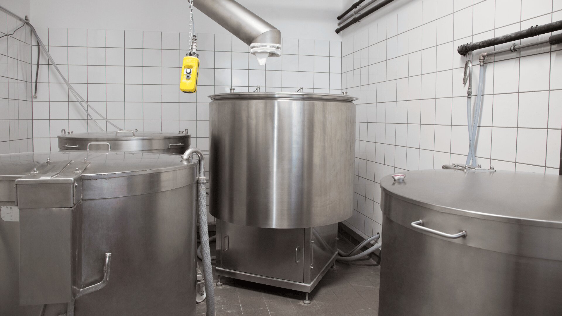 Mehrere große Edelstahltanks mit Deckeln und Rohren sind in einem gekachelten Raum angeordnet, möglicherweise in einer Molkerei oder einem Lebensmittelverarbeitungsbetrieb. Über einem der Tanks hängt ein gelbes Kontrollgerät.