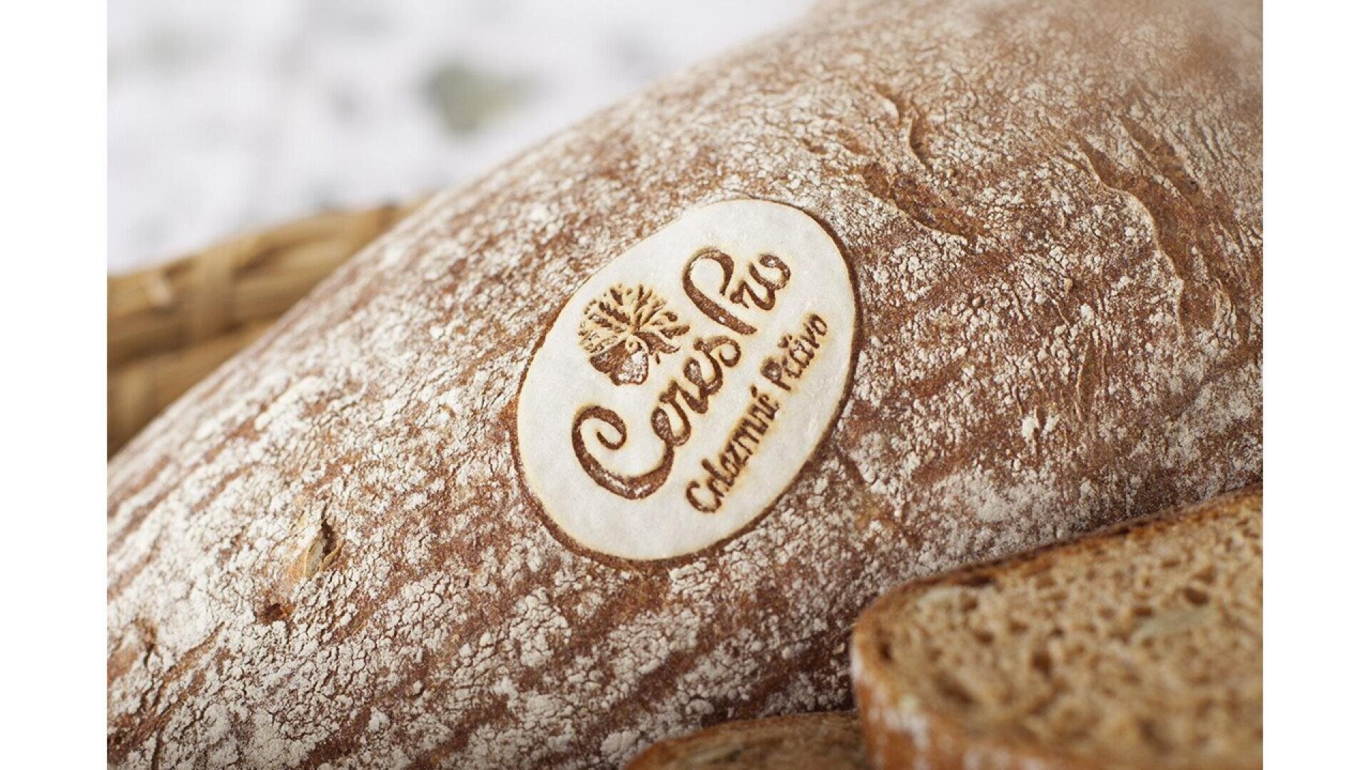 Close-up of a rustic loaf of bread with the CoresRio Colonia Ecovio logo stamped on the crust, next to several slices of bread. The loaf has a dusting of flour on its surface.