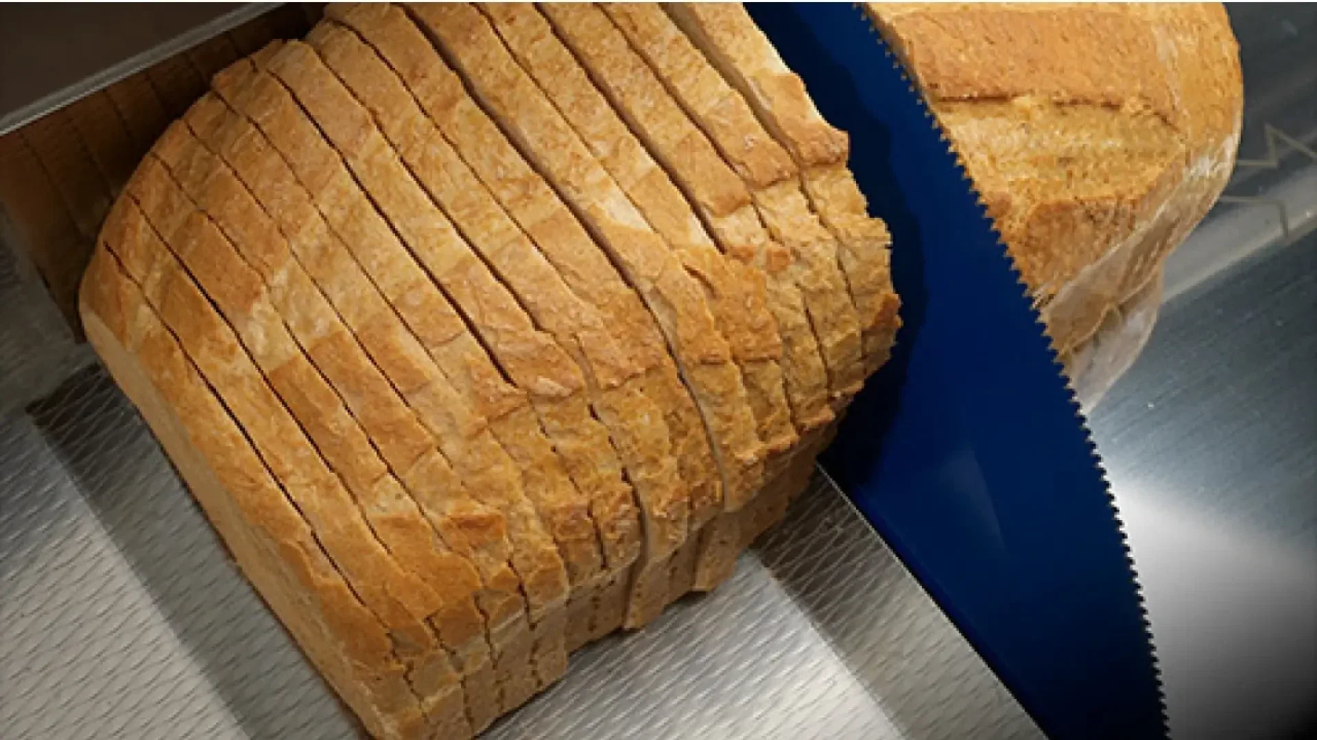 A loaf of bread being sliced into even pieces by a large, blue-bladed bread slicer on a metal surface.