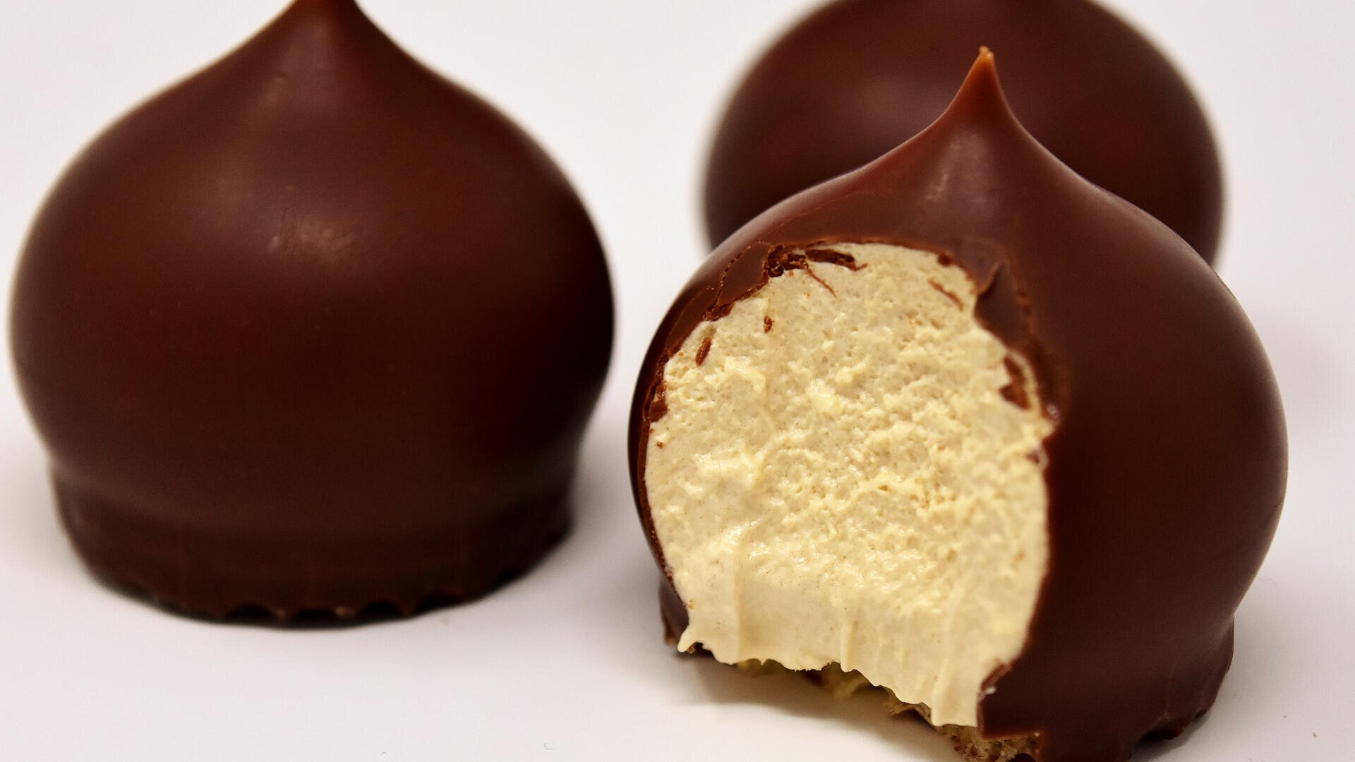 Drei schokoladenüberzogene Bonbons, von denen eines einen Biss hat, der eine cremige, helle Füllung im Inneren offenbart. Die Bonbons sind vor einem schlichten weißen Hintergrund ausgestellt.