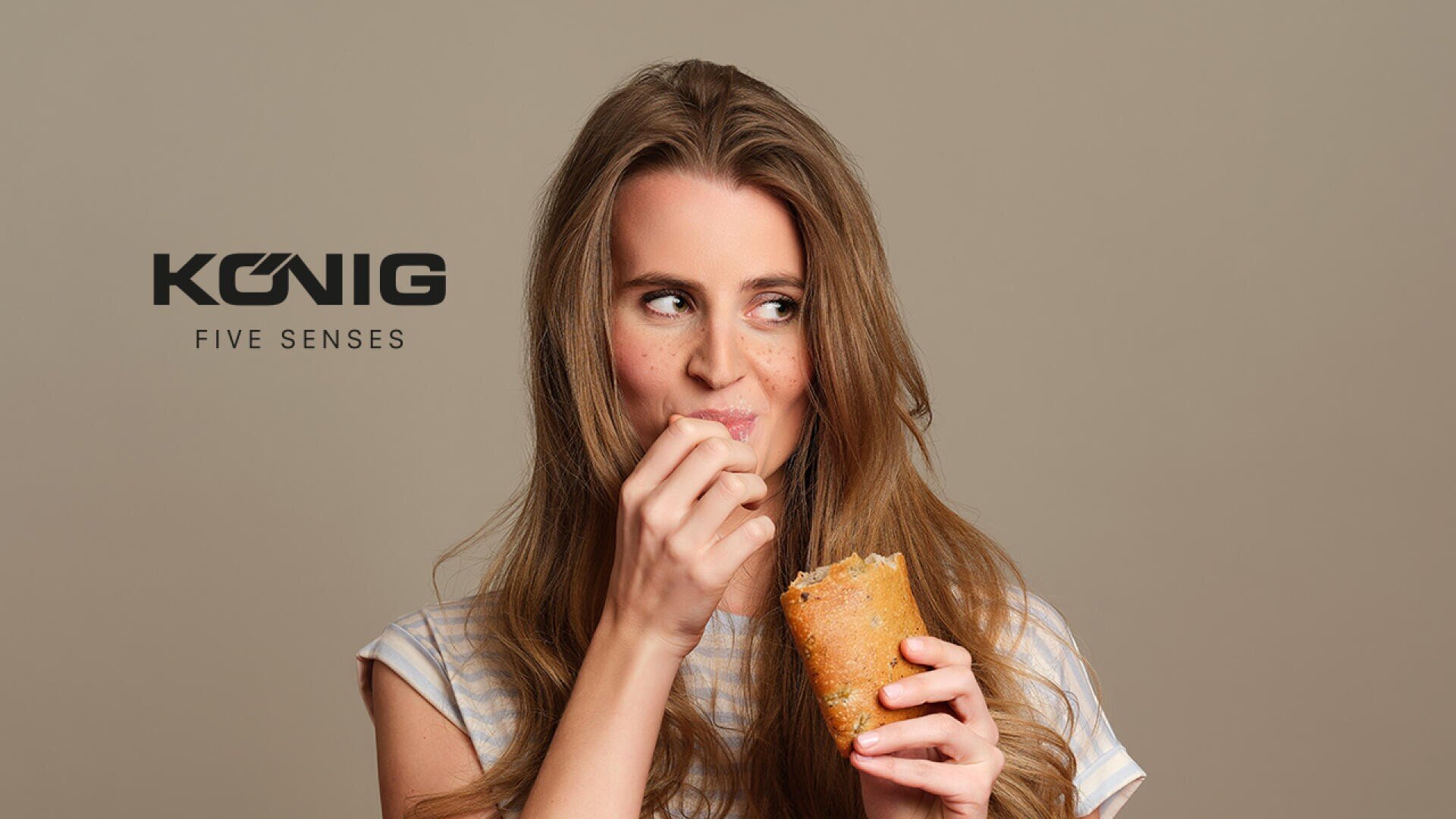 Eine Frau mit langen braunen Haaren lächelt, während sie einen Wrap isst. Sie hält den Wrap in der einen Hand und führt das Essen mit der anderen zum Mund. Der Text KONIG FIVE SENSES erscheint neben ihr auf einem schlichten Hintergrund.