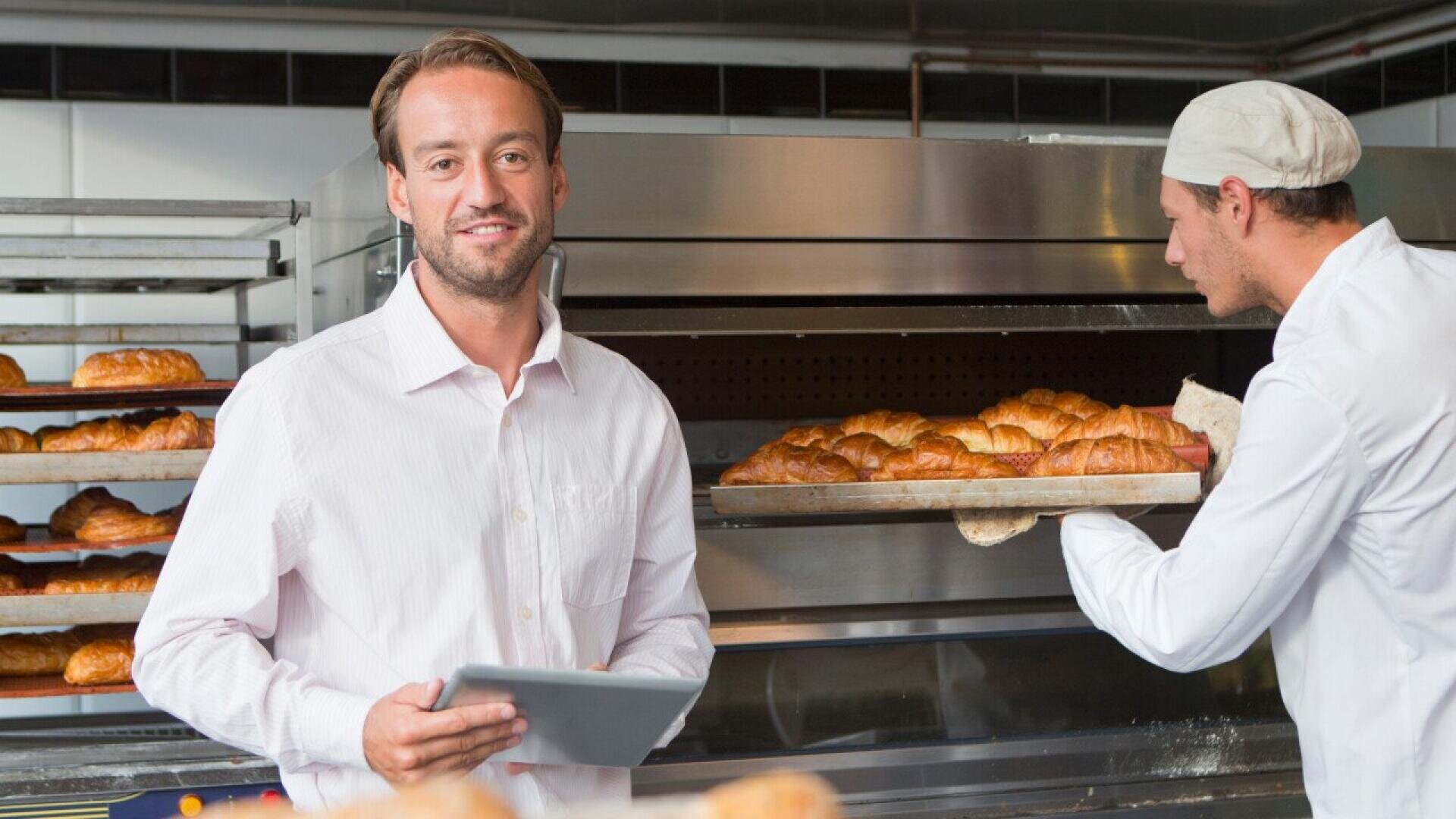 A man in a white shirt holds a tablet and smiles at the camera while another person in bakery attire places a tray of croissants into an industrial oven in a bakery kitchen.