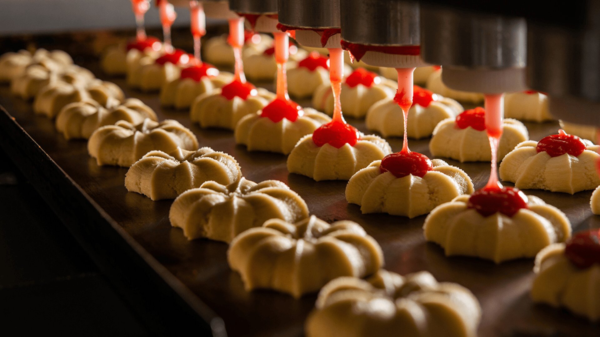 Rows of flower-shaped cookies on a baking tray are topped with red jam by a machine, which dispenses the topping through several nozzles above each cookie in a factory setting.