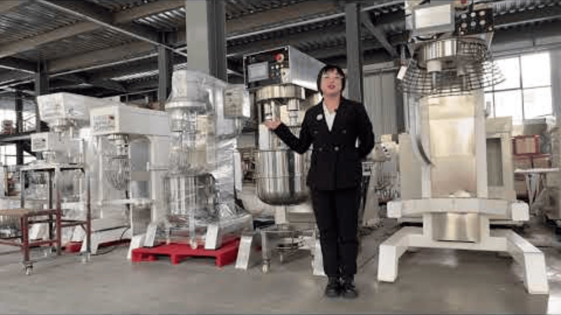 A person in a black suit stands in a factory beside large industrial mixing machines, gesturing toward the equipment. The factory has a high ceiling and metal beams.