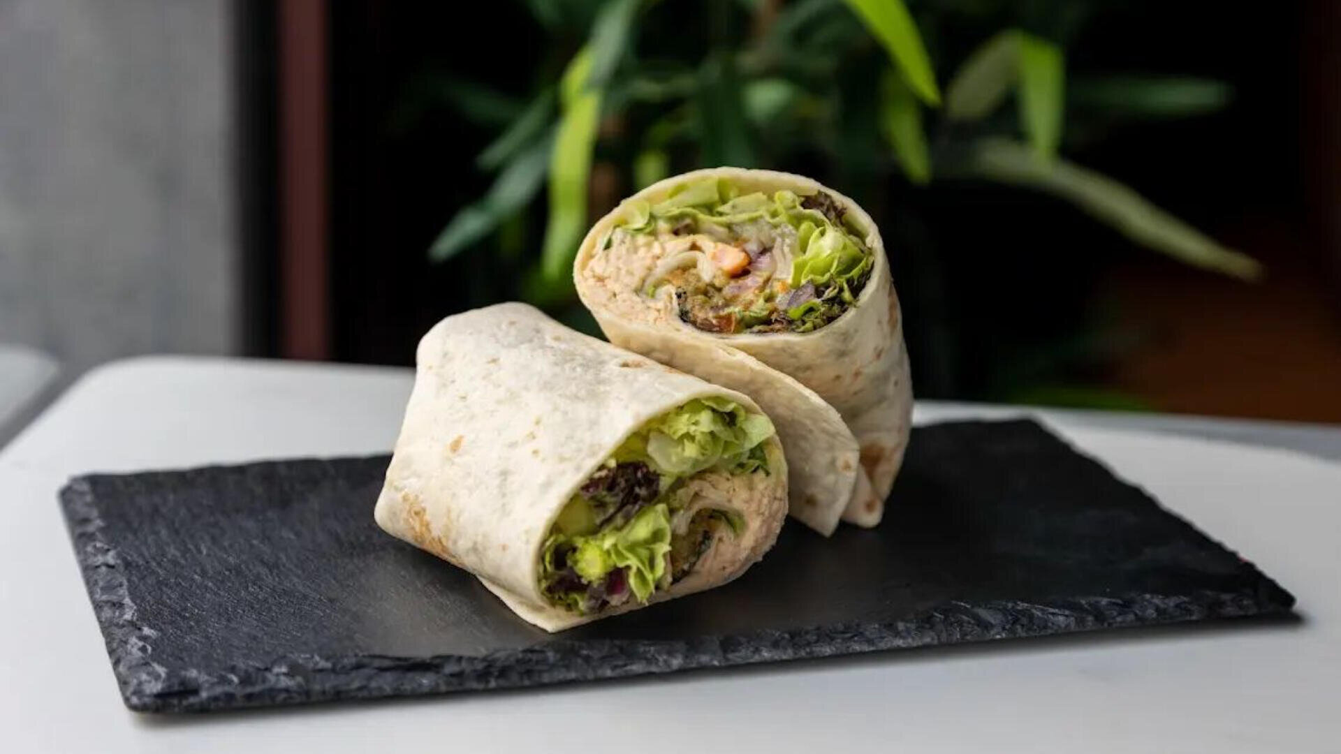 Ein mit Salat, Gemüse und einem cremigen Aufstrich gefüllter Tortilla-Wrap wird halbiert und auf ein rechteckiges Schiefertablett gelegt, wobei eine Blattpflanze im Hintergrund verschwimmt.