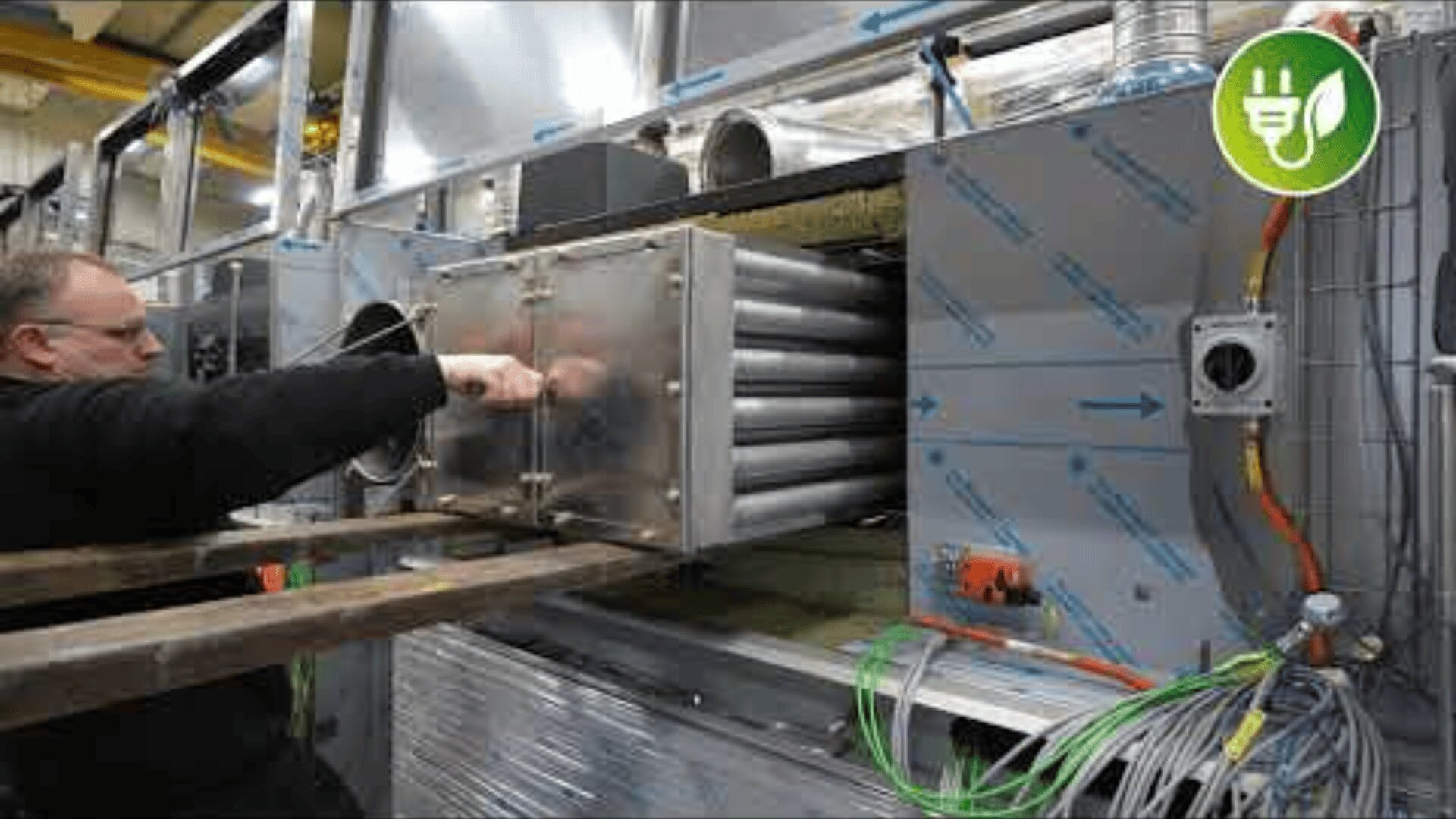 A worker operates or inspects a large industrial machine with exposed metal pipes and wiring, inside a factory setting. A green eco-friendly icon is displayed in the upper right corner.