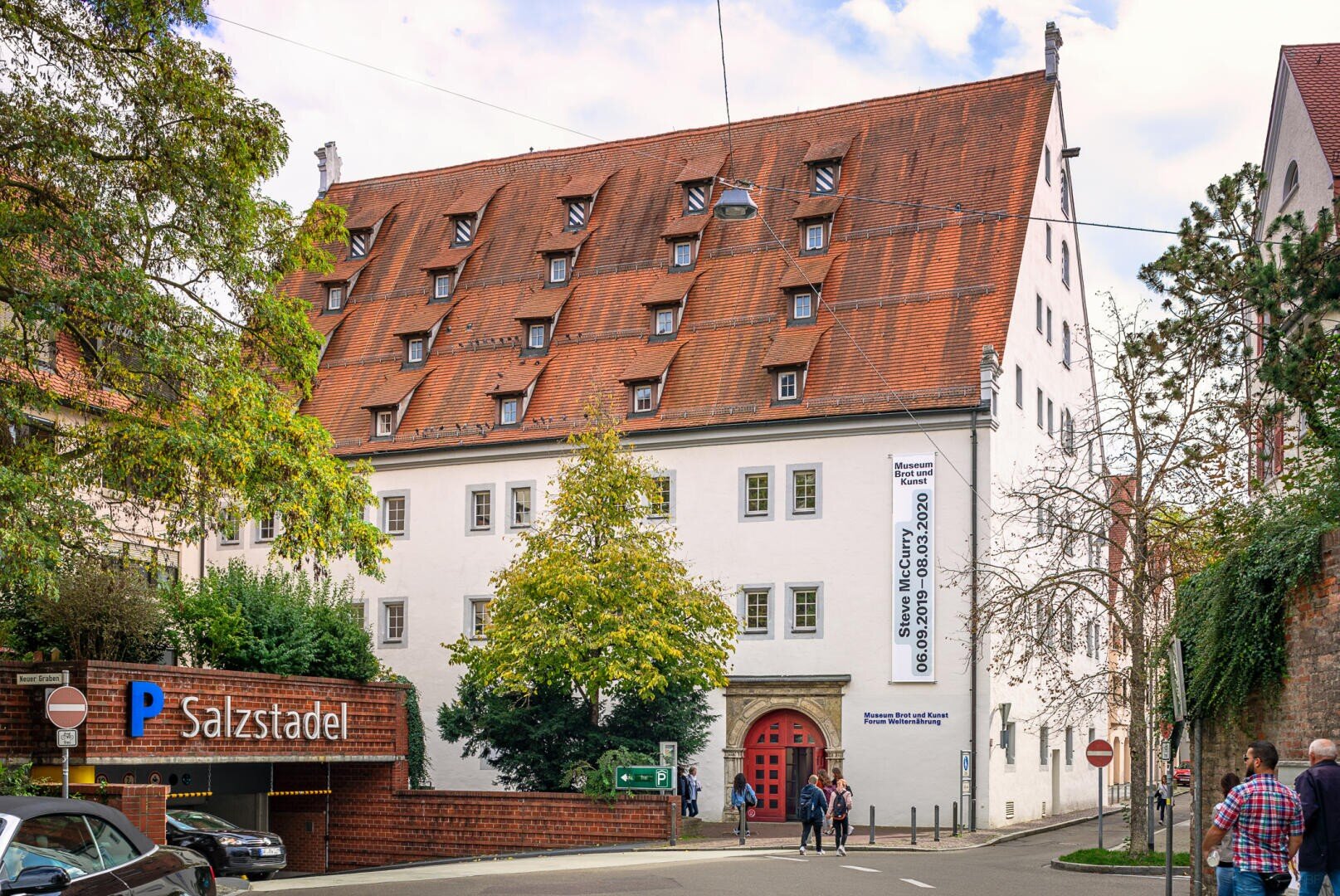 Historisches weißes Gebäude mit steilem roten Ziegeldach und mehreren Dachgauben, das als Salzstadel bezeichnet wird. In der Nähe gehen Menschen spazieren, und es gibt eine Parkeinfahrt mit einem blauen P-Schild. Grüne Bäume und Straßenschilder sind zu sehen.