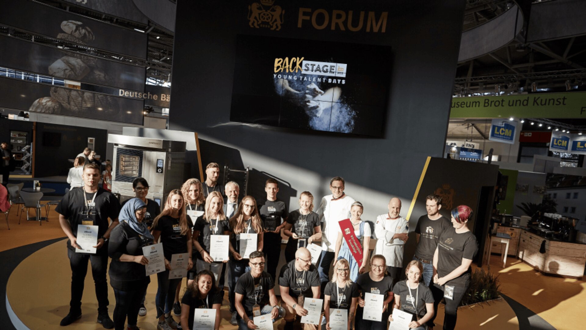 Eine Gruppe von Menschen posiert zusammen mit Urkunden auf einer Bühne unter einem Schild mit der Aufschrift FORUM. Hinter ihnen zeigt ein großer Bildschirm BACKSTAGE YOUNG TALENT DAYS an. Die Szenerie scheint eine Veranstaltung oder Ausstellung zu sein.